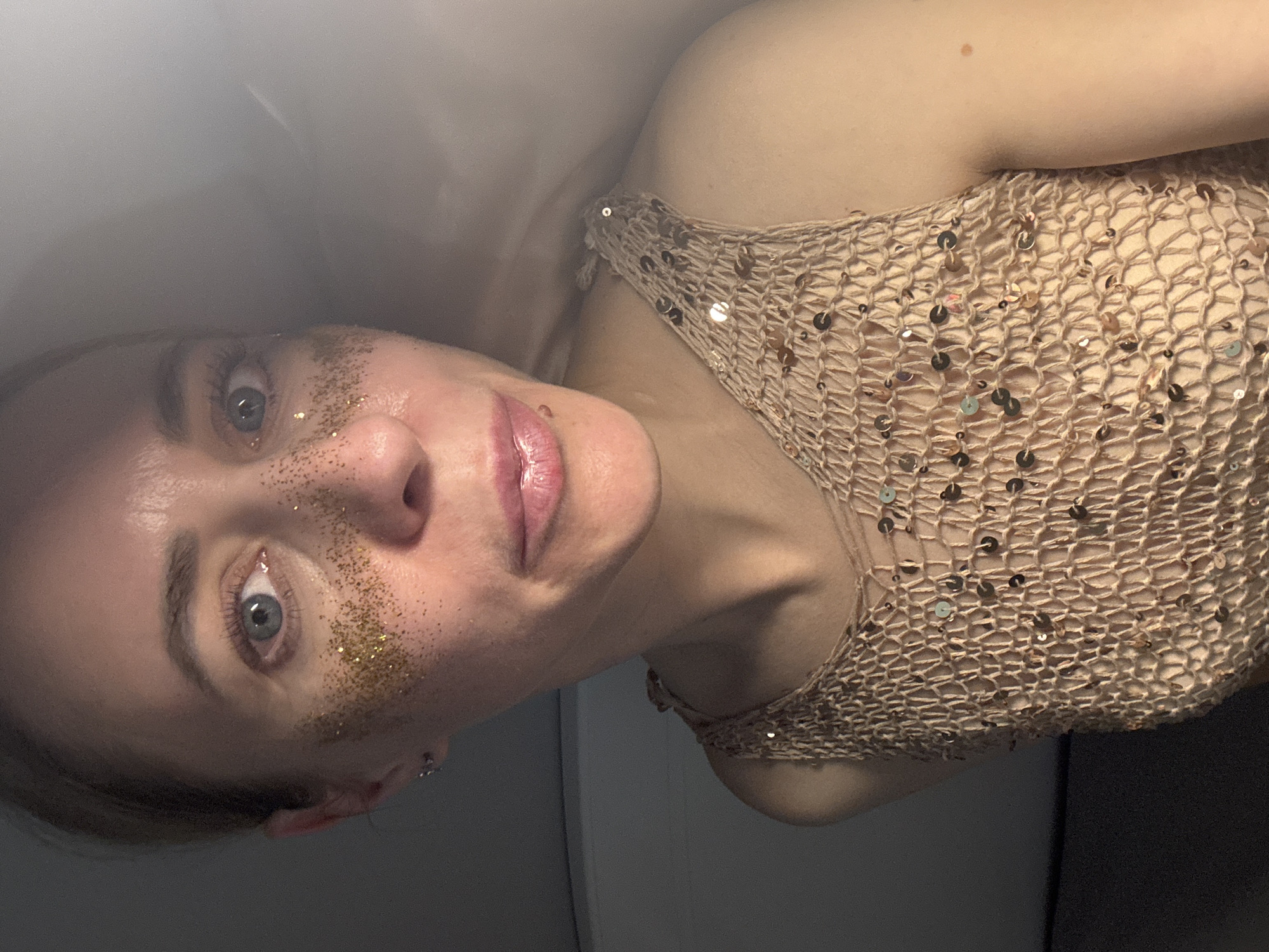 The image displays a close-up portrait of a young adult female in an indoor setting. Her head is tilted approximately 90 degrees clockwise, placing it horizontally on the left side of the frame, with her upper torso extending to the right.
The woman has fair skin, prominent blue-grey eyes, and neatly shaped eyebrows. Her lips are full, appearing glossy and tinted pink. A notable detail is the application of gold glitter on her upper cheeks and beneath her eyes, suggesting a festive, party, or performance-related aesthetic. A small, silver-toned piercing is visible on her lower facial area, possibly near her chin or lower lip.
She is wearing a light beige or tan top made of a loose, open-knit or crocheted fabric, which is embellished with numerous small, reflective sequins in shades of gold and bronze. Her shoulders and upper arm are visible, showing smooth skin.
The background is softly blurred and features muted tones of light grey and darker grey, indicating an indistinct indoor environment with artificial lighting. The woman's gaze is direct, looking towards the camera with a neutral to slightly contemplative expression. No specific location cues are visible within the image itself to confirm the provided context of Chaikovsky, Russia. There is no visible text in the image.