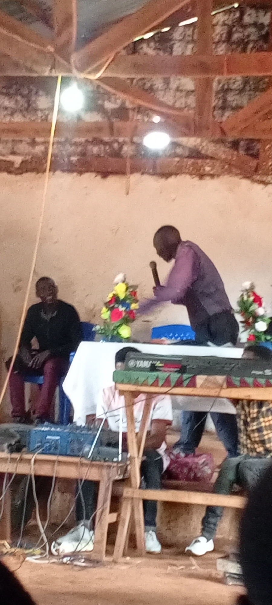 A medium, eye-level indoor shot shows a man speaking into a microphone behind a table, with other people visible at the table and seated on a chair in front of it, possibly in a church setting in Ngalula, Democratic Republic of the Congo. The man speaking is wearing a long-sleeved purple shirt. The table is covered with a white tablecloth, and has flower arrangements on it. Behind the table are people sitting on a wooden structure, some with white shoes on. A keyboard can be seen on top of the wooden structure. Cables are visible on the floor. The walls are rough and unfinished. The ceiling is made of wood beams and a white tarp. There are two lights hanging from the ceiling. A man is seated on a blue chair. He is wearing a black top and burgundy pants.