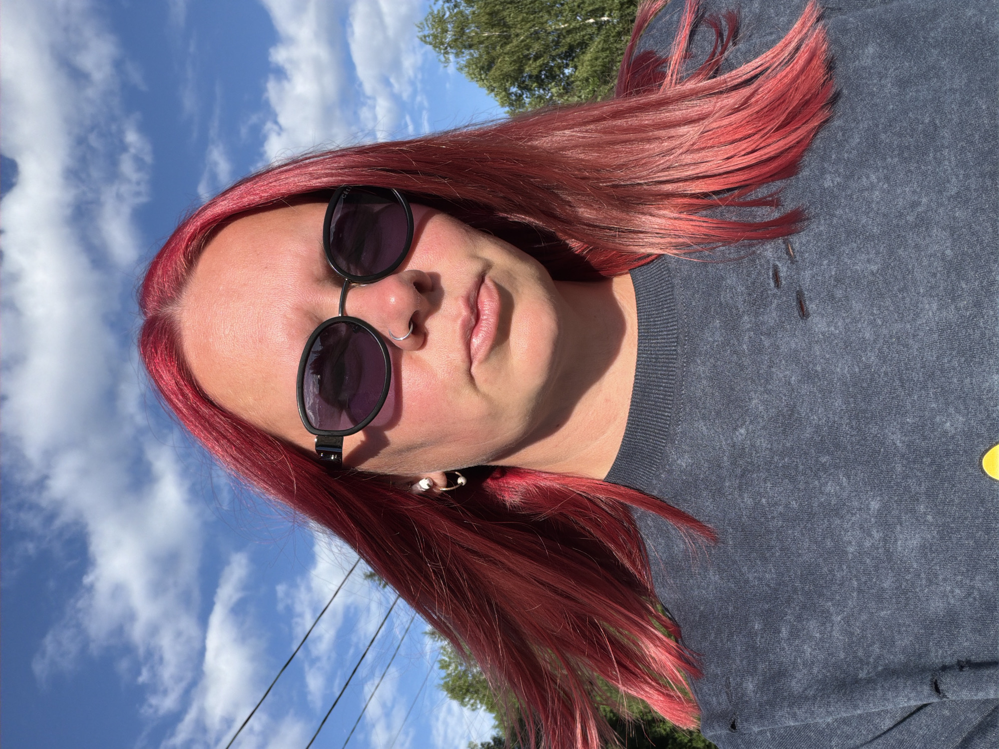 A selfie is taken outdoors under a partly cloudy sky, with power lines visible in the upper left corner and trees in the background. The subject, a person with vibrant red hair and sunglasses, is positioned centrally in the frame, looking directly at the camera. They are wearing a dark, textured crewneck shirt with distressed detailing on the shoulders and chest. A nose ring and hoop earrings are visible accessories. The lighting suggests it is daytime, with shadows cast on the subject's face from the sunglasses and the angle of the sun. The location is identified as Chaikovsky, Russia.