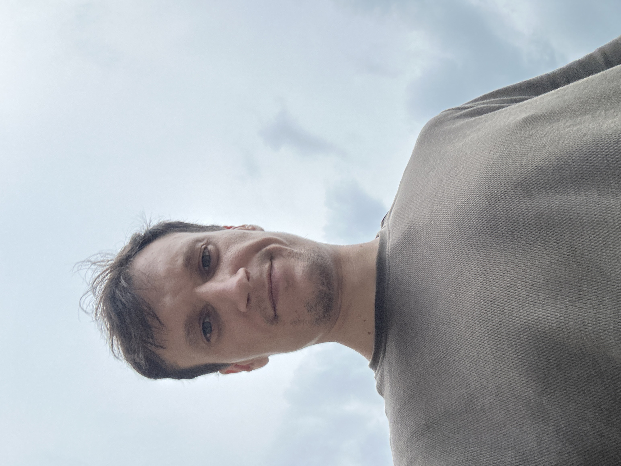 A low-angle, close-up, waist-up selfie of a man with short brown hair and a light beard. He is wearing a brown t-shirt. The background is a pale blue sky with some scattered clouds. The image does not provide any information that would indicate it was taken in Kamianets-Podilskyi, Ukraine.