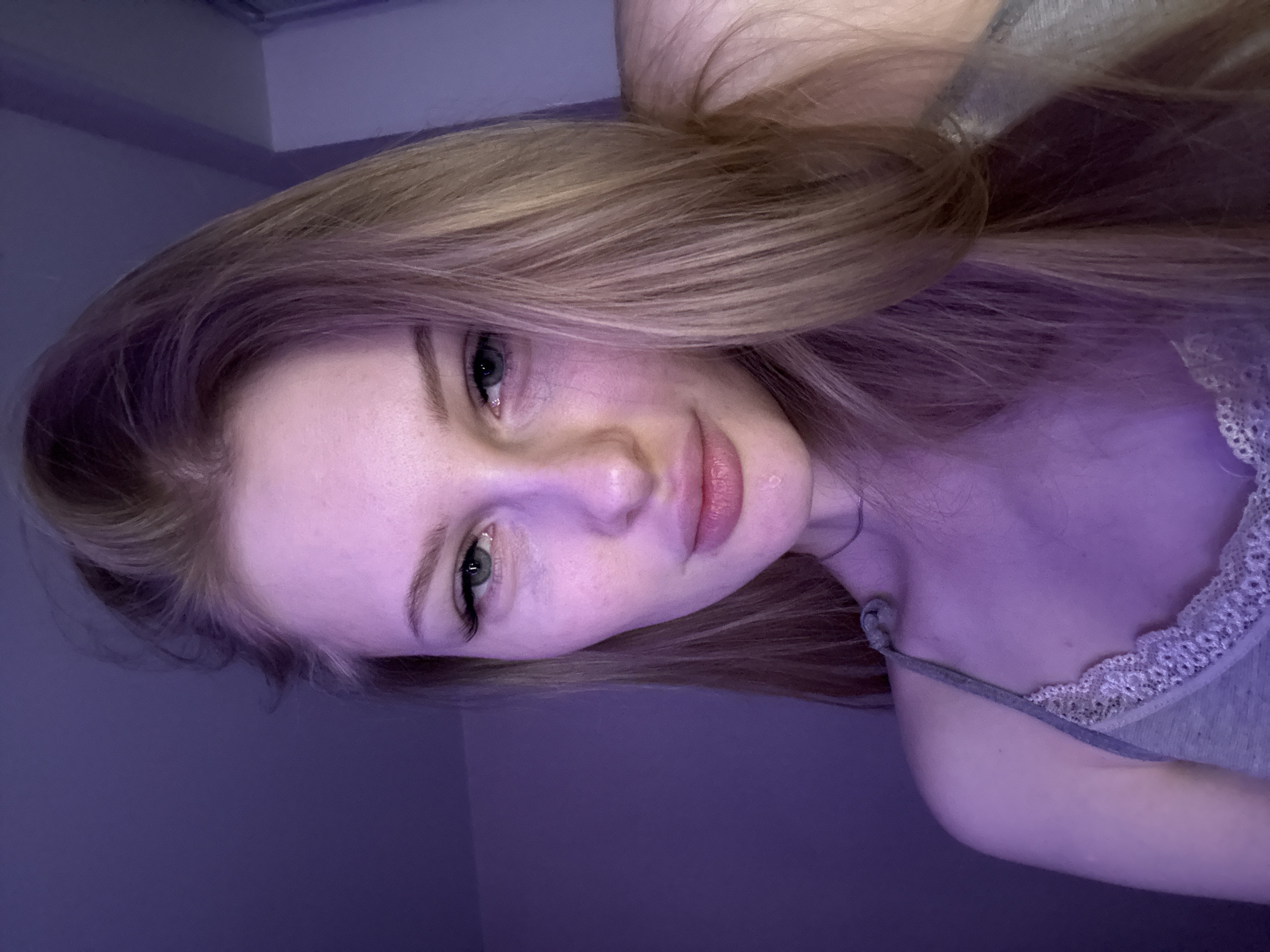 A young woman with light blonde hair, green eyes, and fair skin is facing the camera. She appears to be indoors, possibly in a room with violet lighting that is casting shadows on her face and upper body. She is wearing a gray top with lace trim. The background is a plain wall. There is no explicit indication of Perm, Russia in the image.