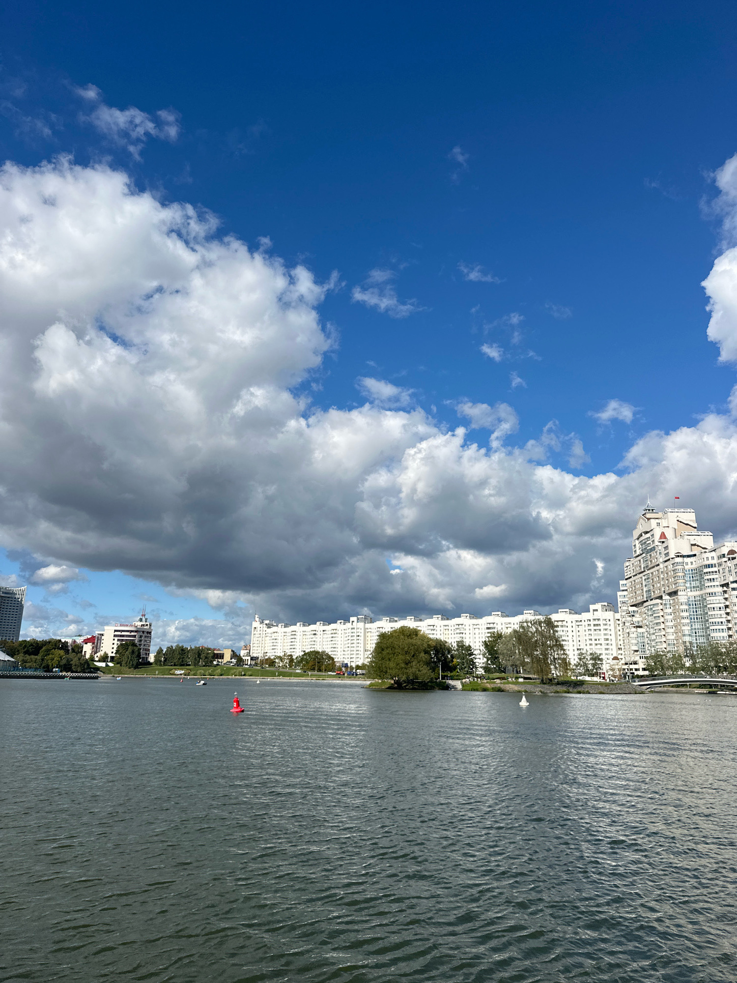 This is a photograph of the Svislach River in Minsk, Belarus, taken on a cloudy day. The sky is a deep blue with large, fluffy white clouds interspersed with darker, more ominous storm clouds. The river is calm, with small ripples on its surface. In the distance, a line of white apartment buildings runs along the embankment, punctuated by trees and a few taller, more modern buildings. There are a few boats on the water, and a red buoy is visible in the foreground. A pedestrian bridge can be seen in the far right of the image. The overall impression is one of urban landscape under a dynamic sky.