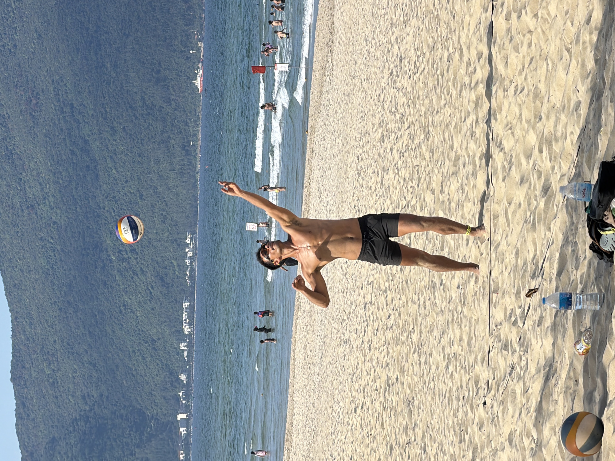 A medium, eye-level shot depicts a man on a sandy beach, appearing to serve a volleyball. He is shirtless, wearing black shorts and sunglasses, and is positioned near a volleyball net. The location is a beach in Đà Nẵng, Vietnam, characterized by a mountain range visible in the background. People are seen in the water along the shoreline, and beach equipment and water bottles are scattered on the sand. The volleyball is multicolored and in motion. The sky is blue.