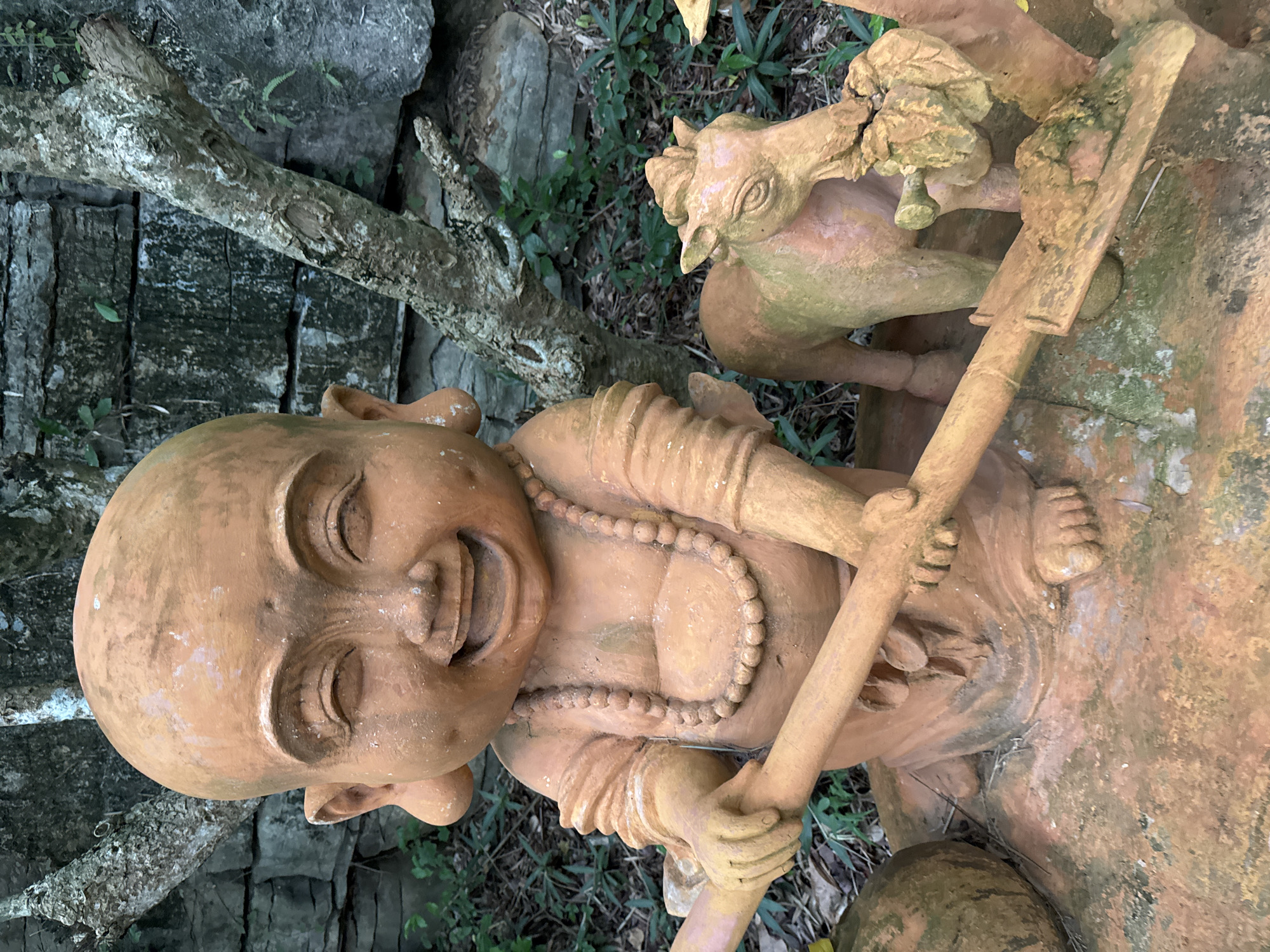 A terra cotta statue of a cheerful, bald-headed child with large ears and a wide grin is the central subject. The child is wearing a beaded necklace and appears to be holding a wooden staff with both hands. To the right of the child is a smaller terra cotta statue of a horse. The statues are situated outdoors, with a stone wall and trees in the background. The ground is covered with dry leaves and some greenery. This statue is located in the historical city of Tây Hoa Lư Ward, Vietnam.
