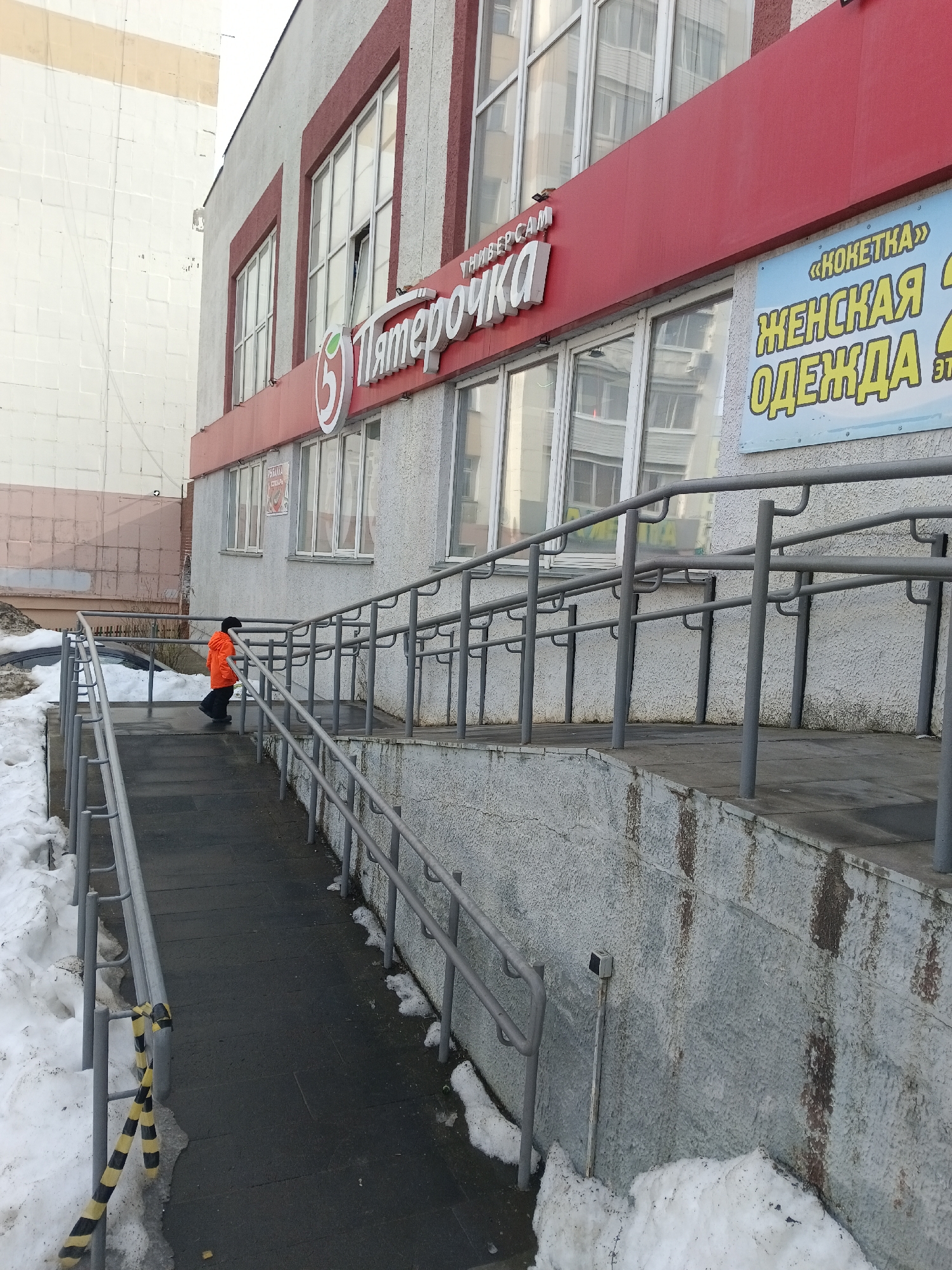 The image depicts an exterior view of a commercial building in Chaikovsky, Russia, during what appears to be late winter or early spring due to the presence of snow. The primary structure is a multi-story building with a light grey, textured facade, accented by red trim around its windows and a prominent red fascia board along the ground floor. A large white sign with a green circular logo on the red fascia identifies the establishment as "Пятерочка" (Pyaterochka), a supermarket, with "УНИВЕРСАМ" (Universal Supermarket) beneath it. To the right of the Pyaterochka sign, a blue sign with yellow text reads "«КОКЕТКА» ЖЕНСКАЯ ОДЕЖДА" (Koketka Women's Clothing).
In the foreground, a dark grey paved access ramp with a gradual incline leads towards the building's entrance, which is not fully visible. The ramp is equipped with continuous grey metal handrails on both sides. Accumulations of dirty, partially melted snow flank the ramp and the building's foundation, indicating cold weather conditions. The lower concrete wall supporting the ramp structure exhibits dark vertical streaks and discoloration. A segment of black and yellow striped caution tape is draped over the left-side handrail near the bottom of the ramp. A dark-colored vehicle is partially obscured by snow and a sparse, leafless bush to the left of the ramp.
One individual, dressed in a bright orange jacket and dark pants, is seen walking uphill on the paved ramp, approximately halfway between the camera and the building entrance. The person's back is to the viewer. Adjacent to the commercial building on the left, a taller, plain white building with visible block patterns is partially visible against a light sky.