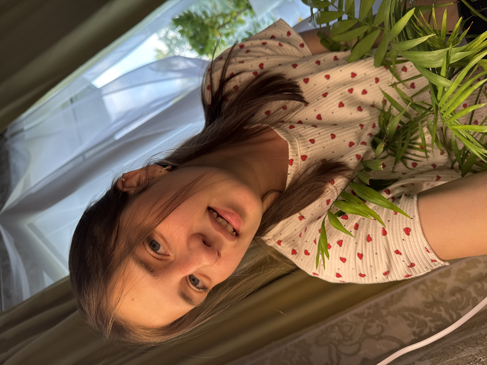 A young woman with light brown hair and blue eyes is pictured from the chest up, looking at the camera with a slight smile. She is wearing a white t-shirt with a pattern of small red hearts. Green leaves from a houseplant are in the foreground, partially obscuring her right side. She has a small earring in her right ear. Behind her, a window is covered by sheer white curtains, and to her left, heavier curtains are visible. The lighting suggests an indoor setting, likely a home. The image does not contain any information about the city of Chaikovsky, Russia.