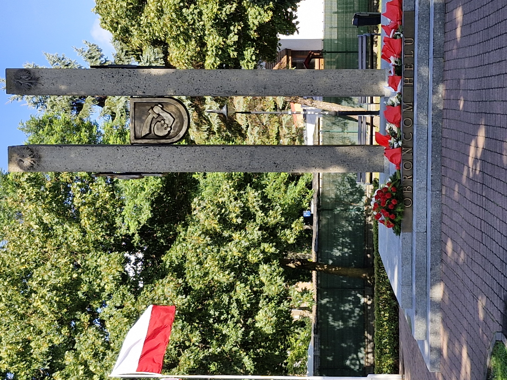 The image shows a monument with two tall concrete pillars, each topped with an eagle emblem. Between the pillars, a coat of arms is displayed, featuring a hand holding a sword within a shield. The monument is situated outdoors, in front of a lush green trees and a clear blue sky. A Polish flag is visible on the left side of the frame, flying from a flagpole. Wreaths of red and white flowers are placed at the base of the monument. The text "OBRONCOM HELU" is inscribed in brass letters on a stone ledge in front of the monument. In the background, there are park benches and a trash can, suggesting a public park or memorial area. The ground in the foreground is paved with brick. The location is identified as the city of Hel, Poland.
