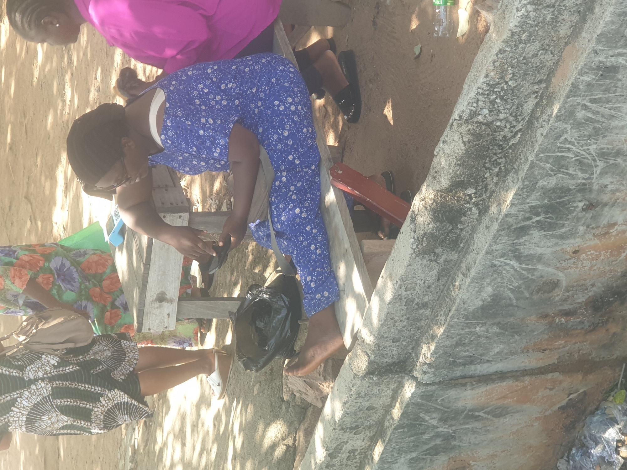 In Jalingo, Nigeria, a group of women are gathered outdoors, seated on and around a wooden bench. One woman, wearing a blue dress with a white floral pattern, is seated on the bench, holding a mobile phone. Another woman, wearing a bright pink garment, is seated to her right. A third woman, in a patterned dress, is standing to the left, with a handbag slung over her shoulder. The ground is sandy, with shadows cast by nearby trees. A black plastic bag is on the ground near the bench. The scene appears to be in an open, possibly informal, public space.