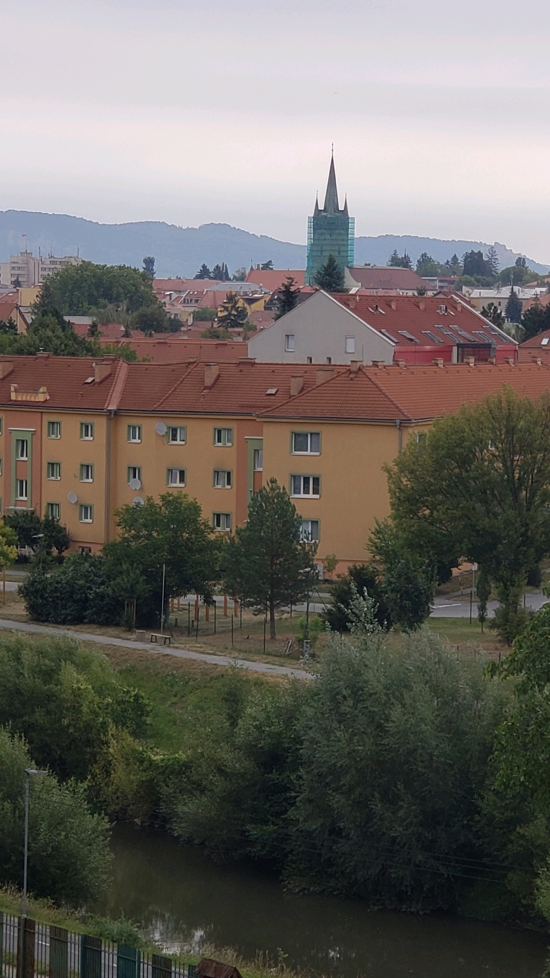 The image captures a view of the city of Prešov, Slovakia, from an elevated perspective. In the background, a prominent green-hued tower with scaffolding, likely a church spire, stands against a hazy, pale sky. Below it, a cluster of buildings with reddish-brown tiled roofs are visible, typical of European urban architecture. In the foreground, a large, mustard-yellow apartment building with multiple windows and satellite dishes is the dominant structure. Adjacent to the apartment building is a strip of green space with trees, a paved pathway, and a solitary bench. Further down, a dark, calm body of water, possibly a river or canal, is bordered by lush, dense vegetation. A black metal fence runs along the bottom edge of the frame, partially obscuring a glimpse of what appears to be a small, brown structure in the lower right corner. The overall atmosphere is overcast, suggesting a cloudy day.