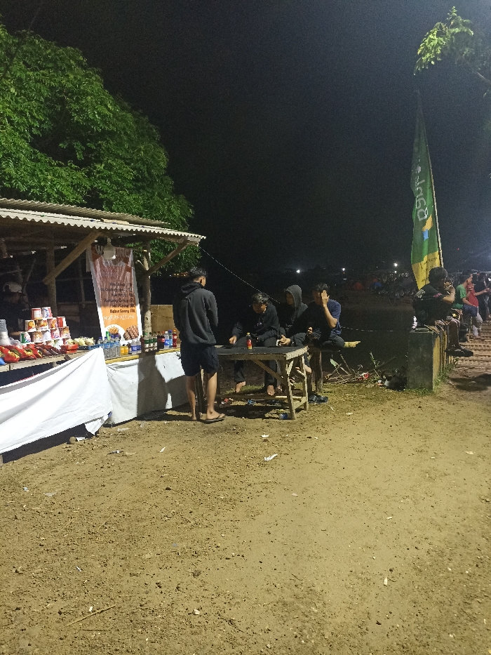 The image depicts an outdoor scene at night in Kecamatan Pujut, Indonesia, centered around a modest roadside stall. The stall features a wooden frame and a corrugated metal roof, illuminated by artificial light sources. It serves as a food and beverage vendor, displaying various packaged snacks, bottled drinks, and canned goods on its shelves. A banner hanging from the stall advertises Indonesian food items, including "JAJANAN" (snacks), "BAKSO" (meatballs), "MIE AYAM" (chicken noodles), "Bihun Goreng" (fried vermicelli), and "MIE AYAM KEJUU" (cheese chicken noodles).
Several men are visible in the foreground. One man in a dark hoodie and shorts stands near the stall, wearing sandals. Three other men, also dressed casually in hoodies, are seated around a low, rustic wooden table, seemingly engaged in conversation or consuming items. Small bottles and cups are on the table. The ground is a rough, unpaved surface.
In the background, a tall green flag with vertical white and yellow text, possibly reading "QSP...", stands near what appears to be a body of water. Distant artificial lights suggest further activity or structures across the water. Another individual is seen sitting on a low concrete ledge by the water's edge. The overall scene suggests a casual, social gathering or a popular local spot operating into the evening hours. The time of day is clearly night, with no visible adverse weather conditions.