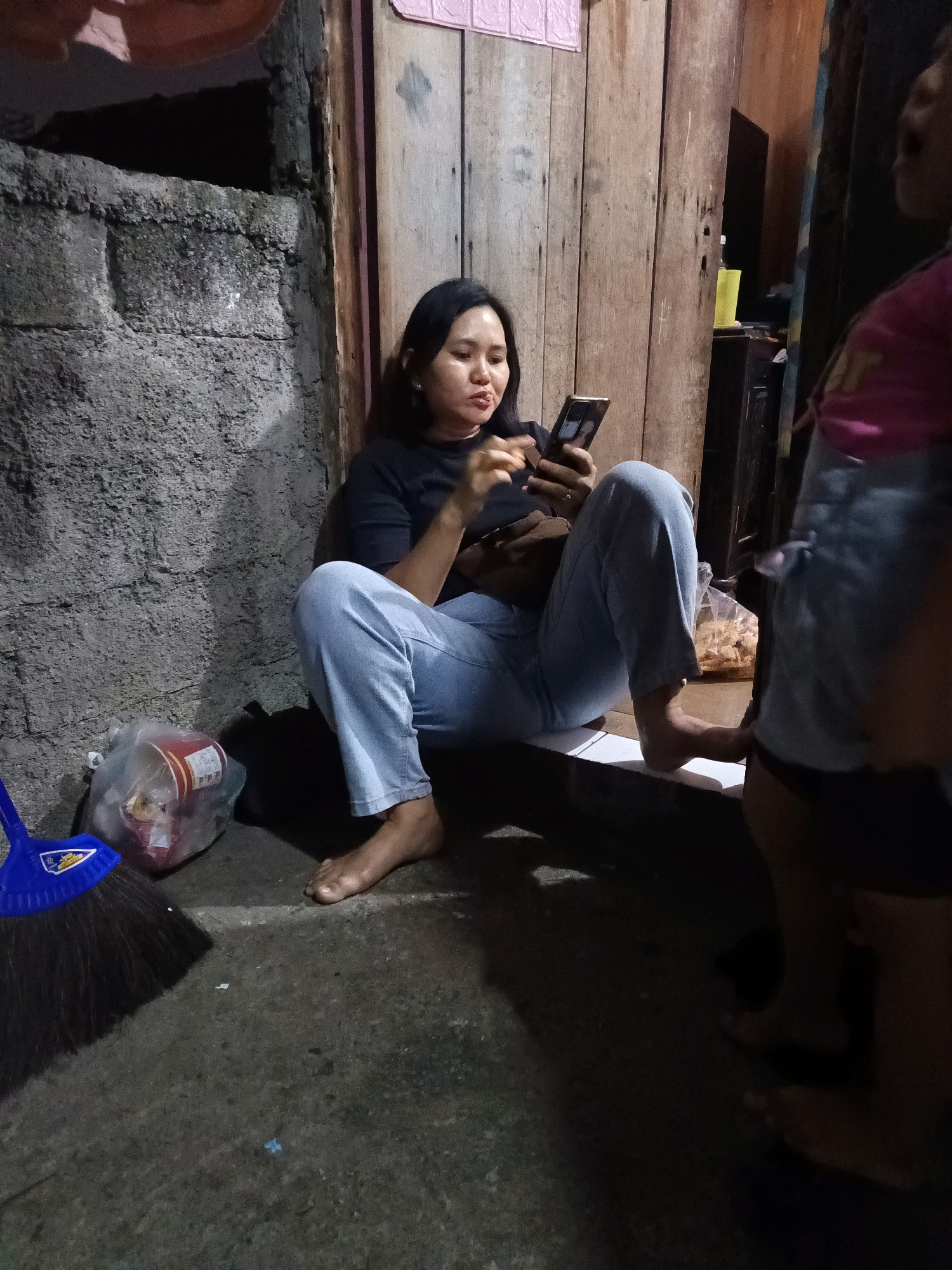 The image depicts an outdoor scene in what appears to be a residential area in South Jakarta, Indonesia, possibly at night or in low light. A woman, dressed in a black t-shirt and light blue jeans, is seated on a step or ledge, looking at her mobile phone. Her posture is relaxed, with her legs crossed and her feet bare. Beside her, on the ground, is a plastic bag containing what appears to be snacks or food items, and a blue broom. In the background, there is a rough concrete wall on the left and a weathered wooden door or wall on the right. Another person, partially visible from the back and in focus on the right side of the frame, is wearing a pink and blue garment, suggesting the presence of at least two individuals. The ground is concrete, and the overall environment suggests a simple, possibly urban, living space.