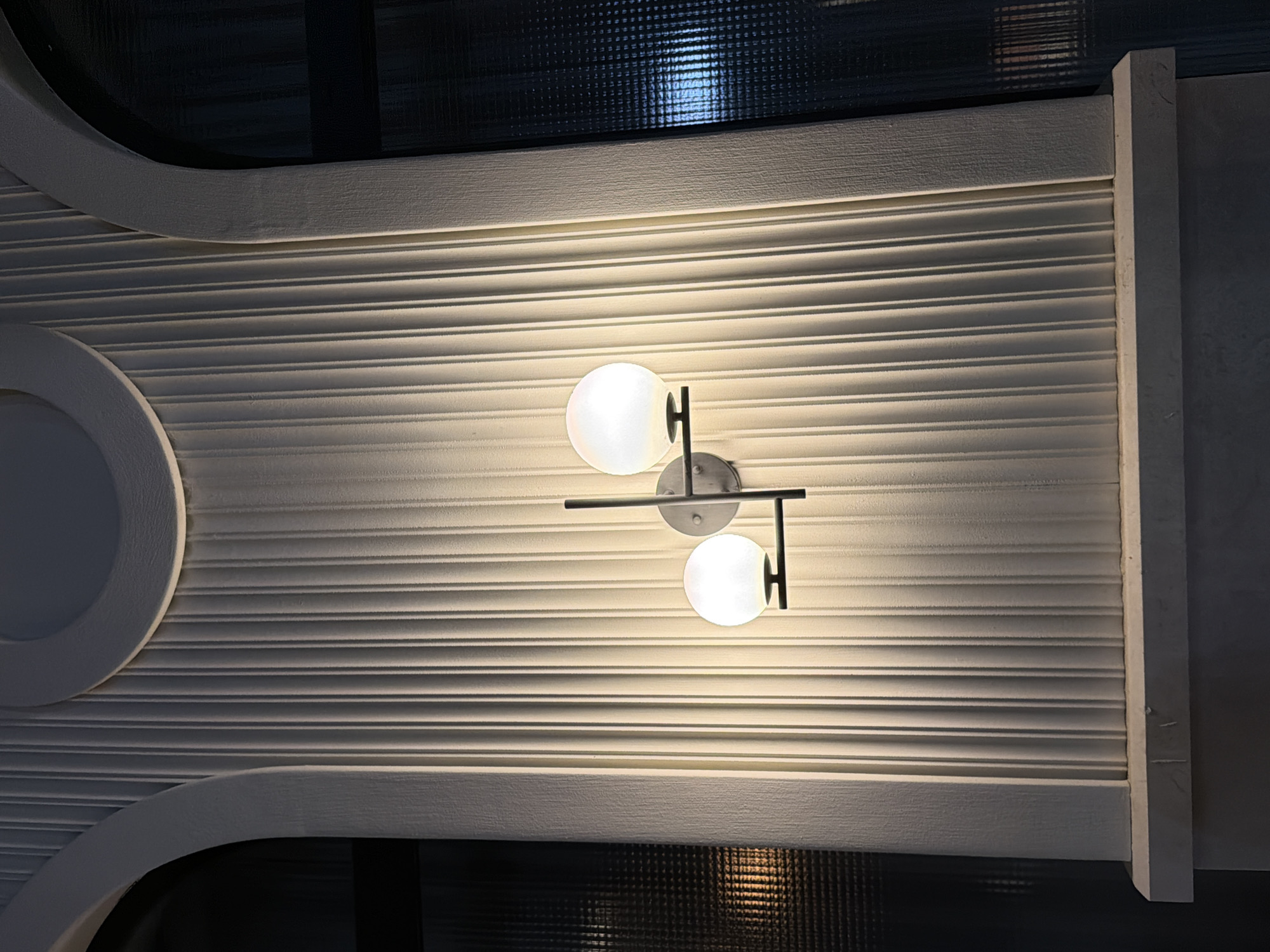 The image shows a close-up of an architectural detail, likely an exterior wall, with a modern wall sconce featuring two spherical lights. The wall is textured with vertical grooves, and arched window frames are visible on either side, with one window showing a dark, textured glass and the other appearing as a black void. The sconce is mounted centrally, casting a warm glow on the ribbed wall. The overall impression is one of contemporary design within a potentially historical or grand setting.
The location is the wall of the Stari Grad fortress in Budva, Montenegro, specifically on the eastern side overlooking the Adriatic Sea.