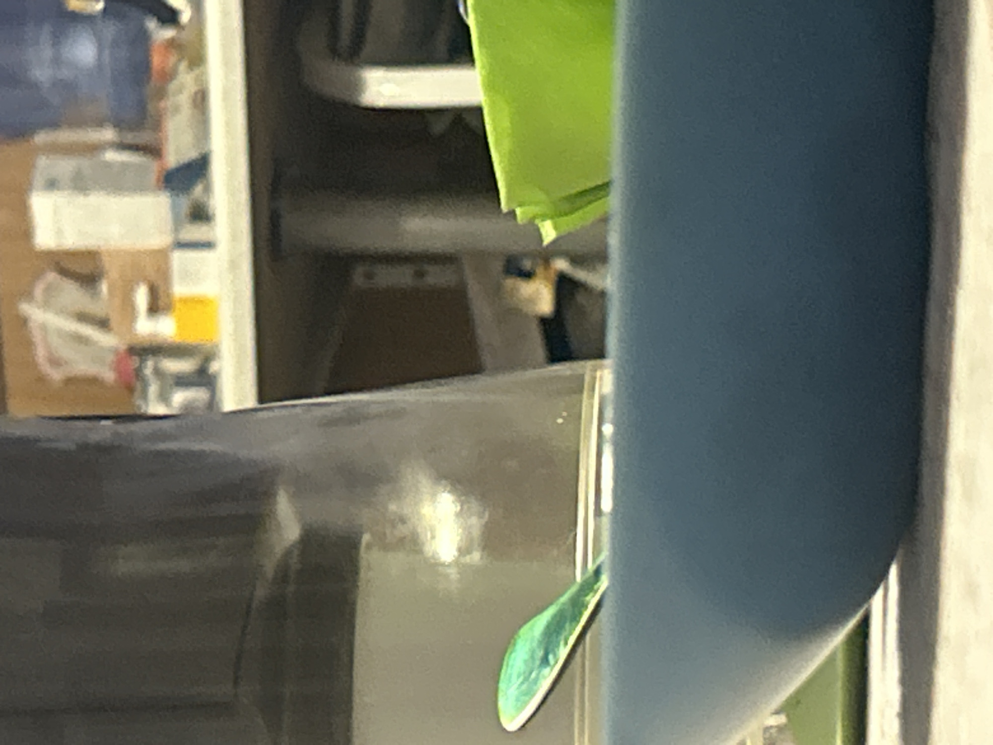 The image shows a close-up of a blue bowl with a metallic green spoon inside. Behind the bowl, a blurry background features a shelf with various items, including a yellow bottle, white boxes, and other indistinct objects. To the right of the bowl, a bright green item, possibly a folded napkin or paper, is visible. The lighting suggests an indoor setting, likely a kitchen or dining area. The overall scene is out of focus, with the bowl and spoon being the most discernible elements. There are no people or clear signs of activity. The location is unknown.