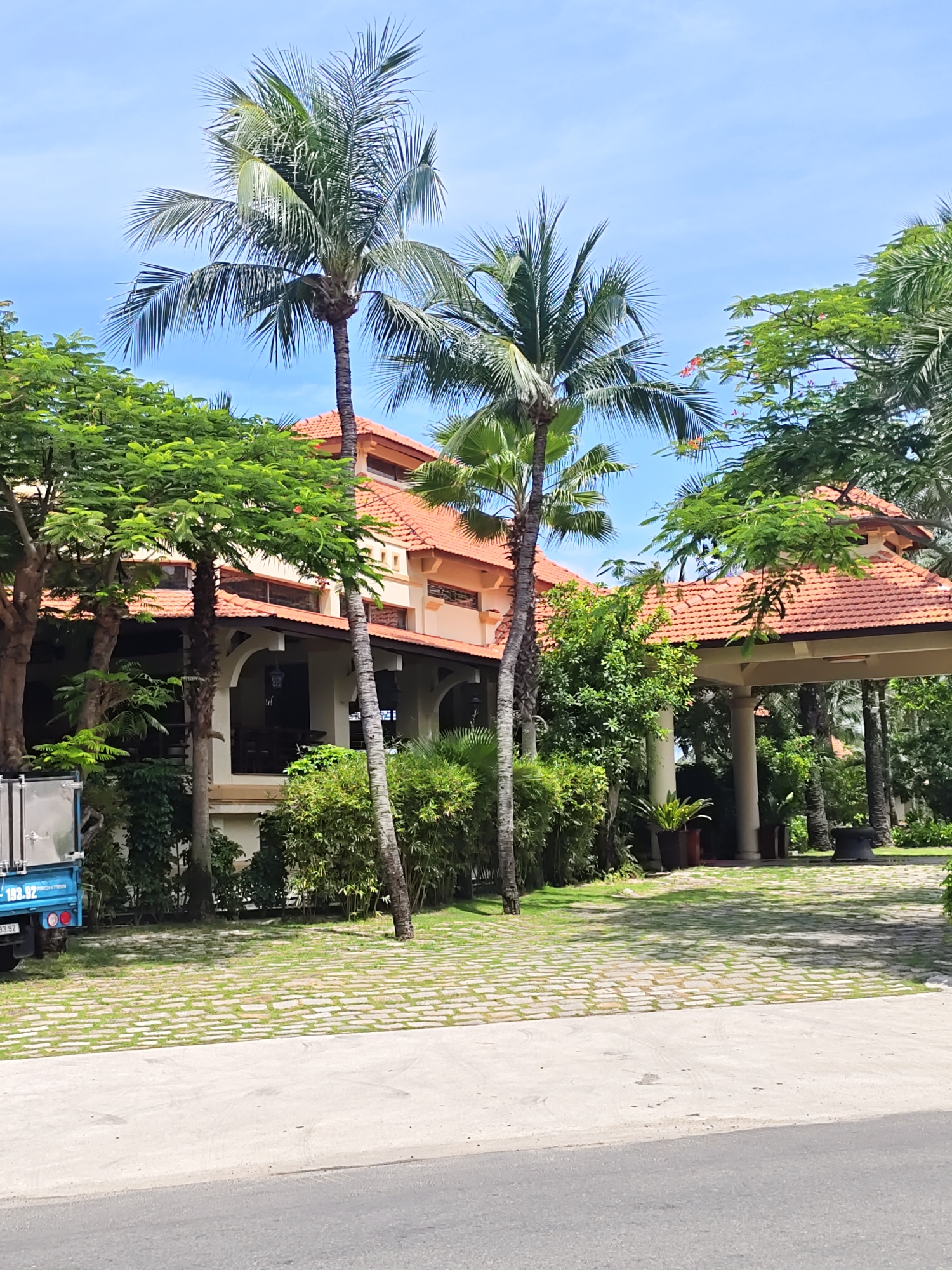 The image shows a resort with a long, single-story building with an orange tiled roof. Palm trees and lush green foliage surround the building. The building has a covered walkway with white pillars. In front of the building, there is a paved area with grass growing between the stones. A blue truck is parked to the left of the frame. The sky is clear and blue.