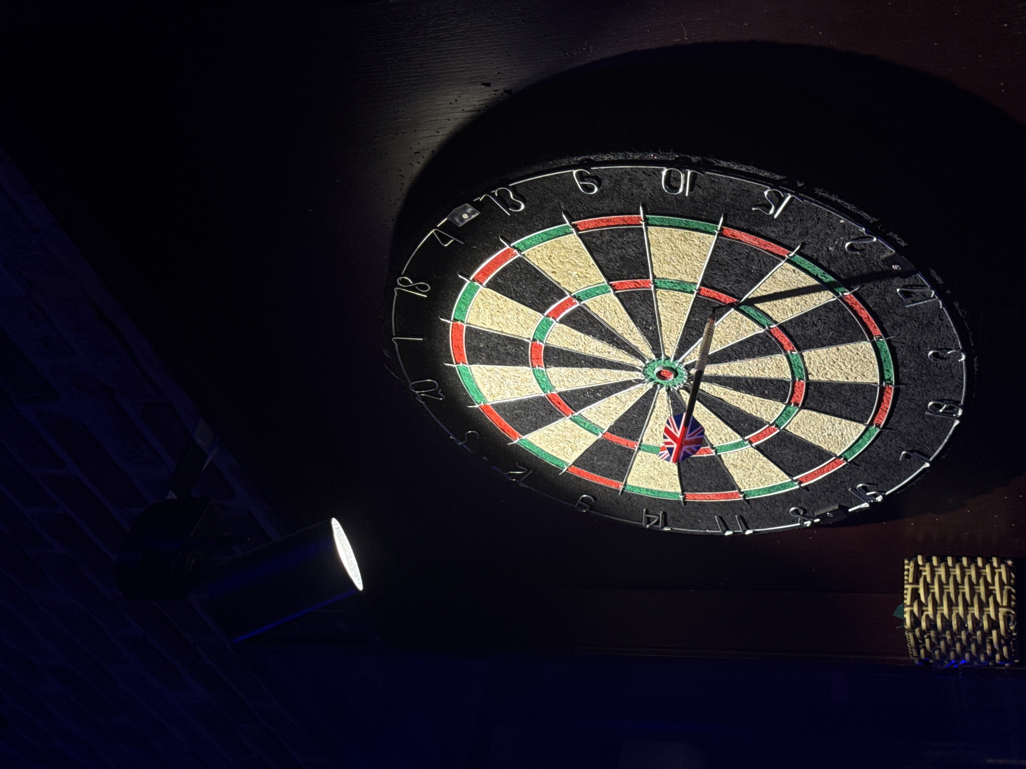 This low-light photograph captures a dartboard mounted on a dark surface, likely a wall, in Đà Nẵng, Vietnam. The dartboard is the central focus, dramatically illuminated by a bright spotlight positioned to its lower left. The rest of the scene recedes into deep shadow, emphasizing the circular target.
A single dart, adorned with a Union Jack flag on its flight, is stuck in the dartboard near the bullseye, suggesting that a game or practice session has recently taken place or is in progress. The dartboard displays its standard numbering system around its perimeter, with visible numbers including 18, 4, 13, 6, 10, 15, 2, 7, 19, 3, 17, 8, 16, 11, 14, 9, 12, 5, 20, and 1.
To the lower right of the dartboard, a small, woven, possibly rattan, basket or holder is partially visible. The artificial spotlight casting a strong, directed light onto the dartboard indicates an indoor setting, such as a pub, game room, or private recreational area, and suggests the time of day is evening or night. No people are visible, and no specific weather conditions are discernible from the indoor scene. The overall atmosphere is focused and dark, highlighting the game itself.
