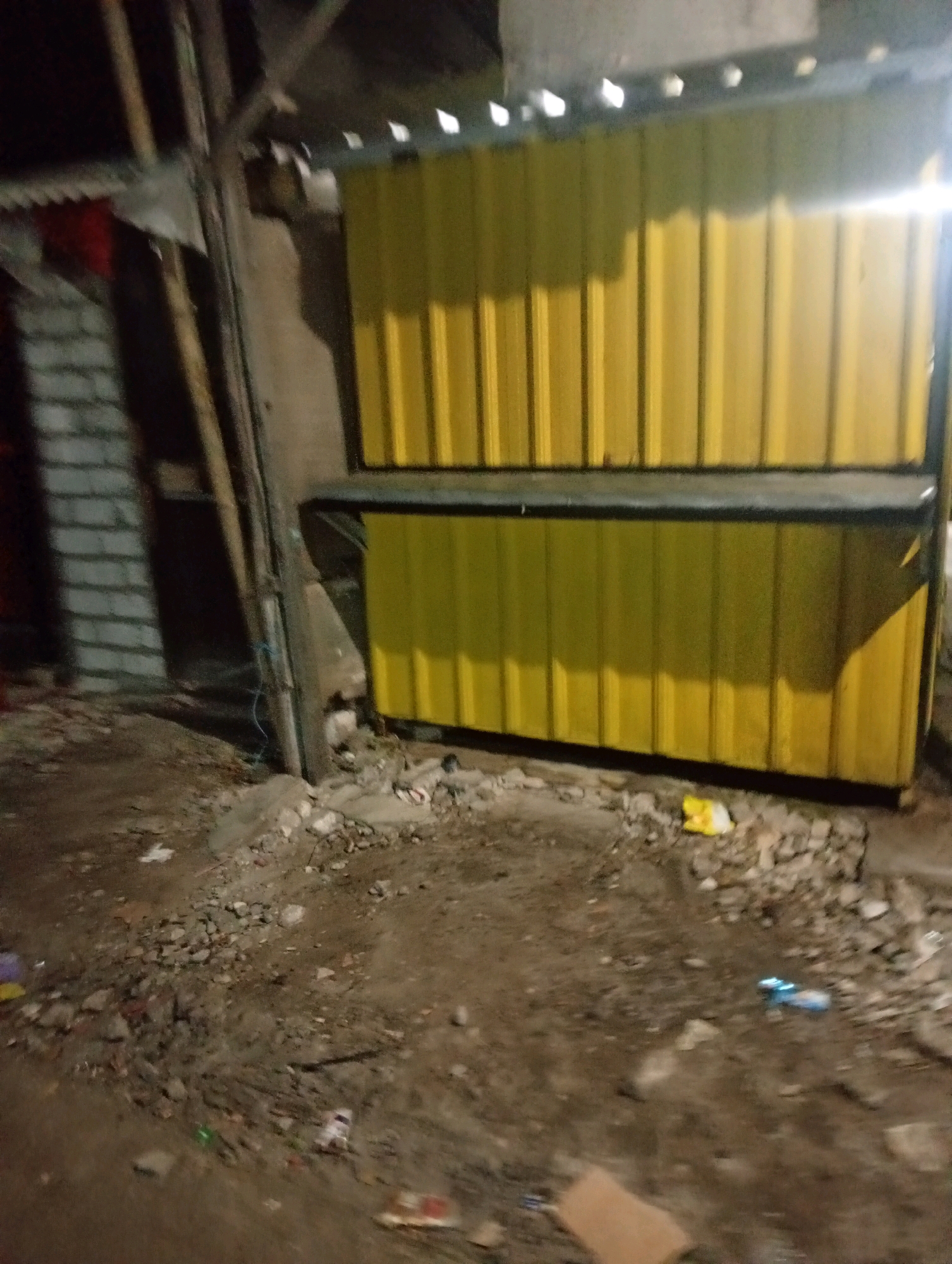 The image depicts an outdoor scene at night, likely in Banowan, Indonesia, characterized by a somewhat cluttered and unkempt environment. The focal point is a structure with two stacked yellow corrugated metal panels. The lower panel is directly accessible from the ground, while the upper panel is raised, supported by dark vertical beams. A dark horizontal band separates the two yellow panels. To the left of the structure, a brick wall with a visible pattern is partially illuminated, and a vertical wooden pole is positioned in front of it. The ground is uneven, covered with dirt, small rocks, and scattered debris, including what appears to be discarded plastic packaging, a crumpled yellow bag, and other unidentifiable items. Lighting conditions suggest artificial illumination from an unseen source, casting shadows on the yellow panels. The overall impression is of an outdoor storage or workshop area that is not meticulously maintained.