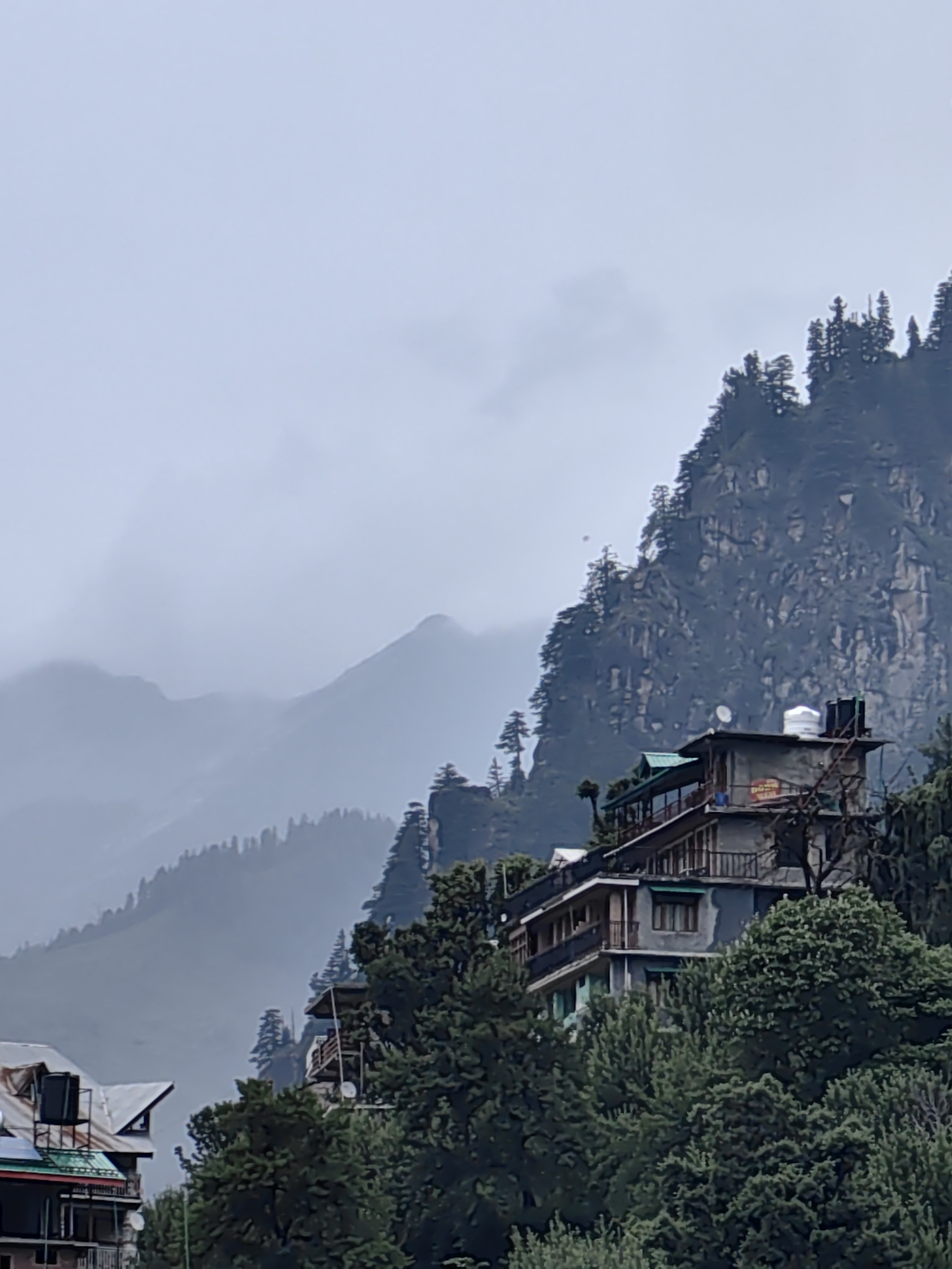 This is a vertical, medium-shot photograph of a building perched on a steep, forested mountainside in Vashisht, India. The building appears to be a guesthouse or hotel, with multiple levels, balconies, and wooden railings. It is constructed with concrete and has a dark grey exterior. Green awnings are visible over some windows. On the rooftop, there are two water tanks, one white and one black, and some plumbing. A circular sign with red and yellow text is attached to the side of the building, but the text is illegible due to the distance and image resolution.
The building is nestled amongst dense, dark green trees that cover the lower slopes of the mountain. Behind and above the building, the mountain rises steeply, characterized by rugged rock faces and sparse vegetation, primarily coniferous trees. The upper parts of the mountains are shrouded in mist and clouds, suggesting a hazy or rainy day, possibly in the early morning or late afternoon given the diffused light. In the far background, successive layers of mountains recede into the distance, fading into a pale, overcast sky. The atmosphere is serene and somewhat dramatic, with the imposing natural landscape contrasting with the man-made structure. A smaller building with a metal roof is partially visible in the lower left corner of the frame.