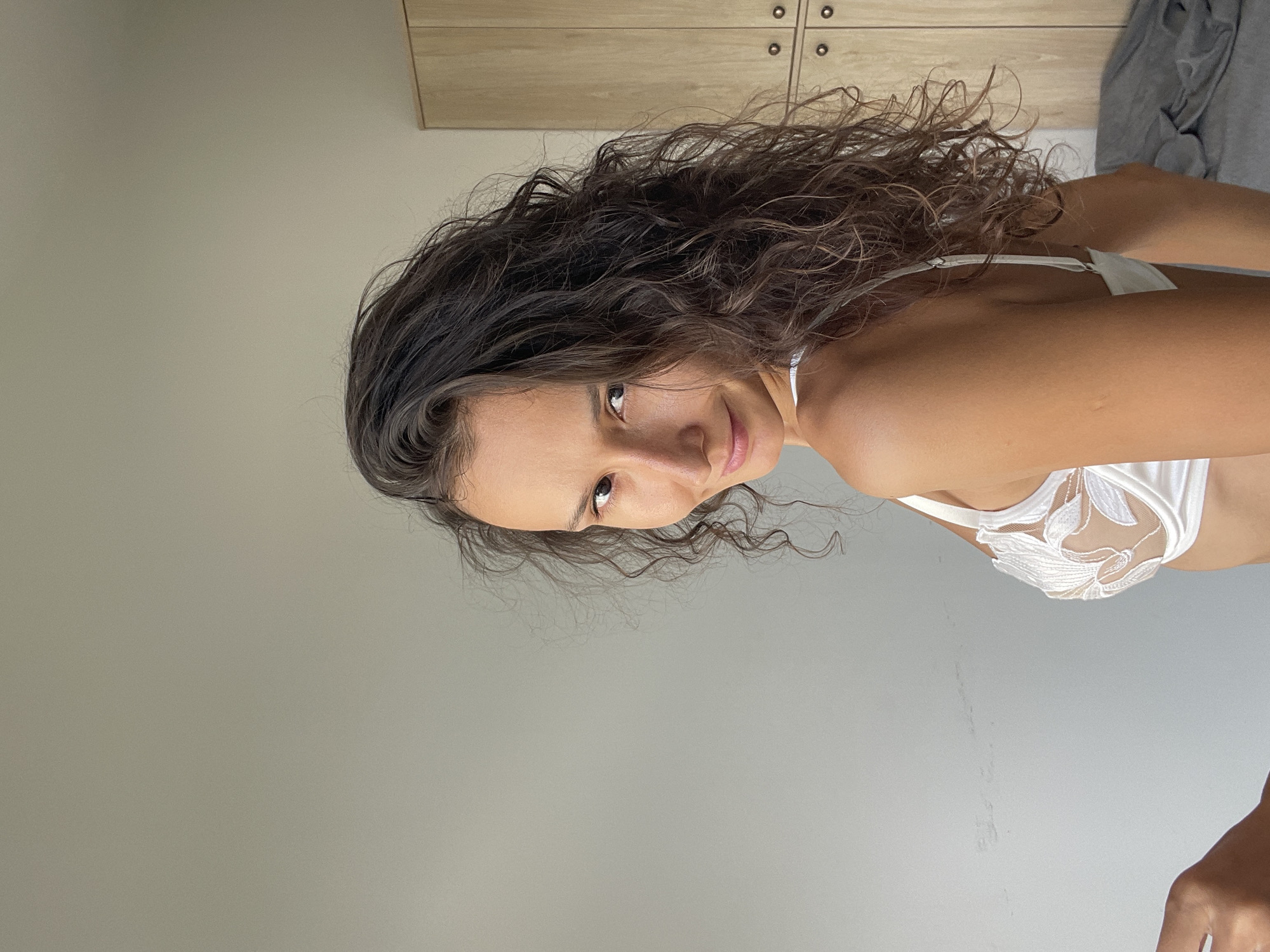 A woman with dark, curly hair and light skin is shown from the chest up, looking over her shoulder at the camera. She is wearing a white bra with intricate floral lace detailing. Her skin appears tanned. The background is a plain, light-colored wall with a wooden cabinet to her right. The scene appears to be indoors, possibly a bedroom. The image is cropped to focus on the woman. There is no indication of Phu Thuy Ward, Vietnam, or any other specific city or location within the image or its provided crops.