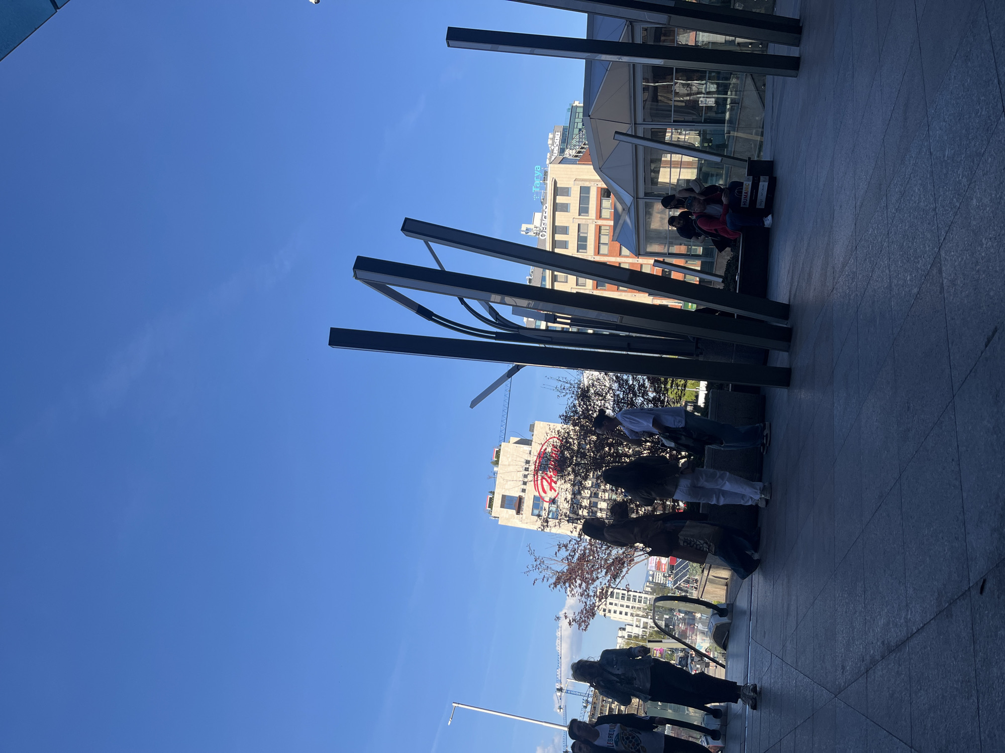 The image depicts a street scene in Sofia, Bulgaria. The sky is a clear blue. In the foreground, several metal beams form an architectural structure. People are present in the image, some standing and walking along a paved walkway, others sitting by a donation bin on a bench. There are buildings in the background, including one with a prominent red logo resembling a circle divided into sections. The building is the Bulgarian American Credit Bank (BACB) at 2 St. Sofia, Bulgaria.