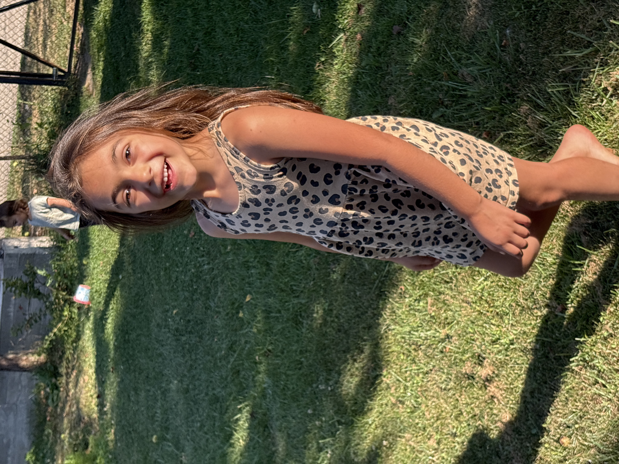 A young girl with long brown hair and a leopard-print dress is smiling at the camera, her legs crossed. She is outdoors on a grassy lawn, with dappled sunlight filtering through trees, suggesting it is daytime. In the background, another child is visible, bending over, and a fence with a metal frame. A small, red and white bucket is on the ground near a bush on the left side of the frame. The scene appears to be a casual, outdoor setting, possibly a backyard or park. The girl's expression is happy and joyful. The location is identified as Oreshak, Bulgaria.