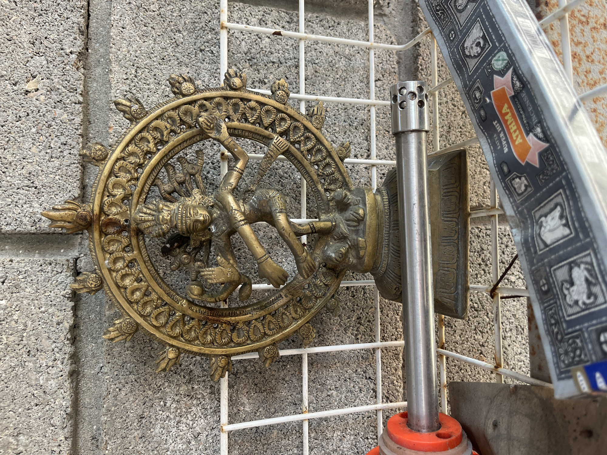 A brass statue of the Hindu deity Shiva as Nataraja, the lord of dance, is displayed against a rough, grey cinder block wall. The statue, which depicts Shiva in his cosmic dance pose within a flaming halo, is supported by a circular pedestal. In front of the statue, a metal pipe with an orange attachment and a box of "ATHMA SATYA SUPER HIT INCENSE" are placed on a white wire rack. The overall scene appears to be an outdoor setting, possibly a market or a decorative display area.