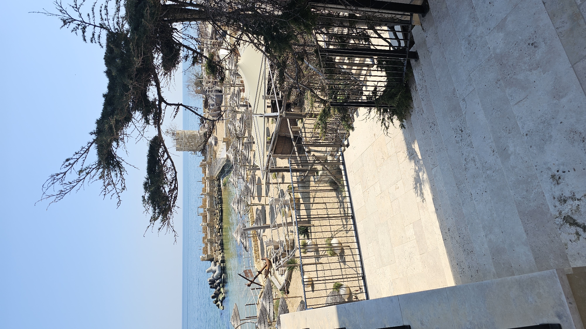 The image shows a beach scene in Varna, Bulgaria, from an elevated perspective. The sky is clear and blue. A pine tree hangs over the scene from the upper left corner. Below, there is a beach with a mix of sand and water, lined with wooden breakwaters and tetrapods. There are lounge chairs and umbrellas on the beach, suggesting a resort area. The water is calm and light blue. In the foreground is a stone patio with a metal railing. To the right of the frame are stone steps, indicating a pathway. The scene appears to be taken from a hotel or restaurant overlooking the beach.