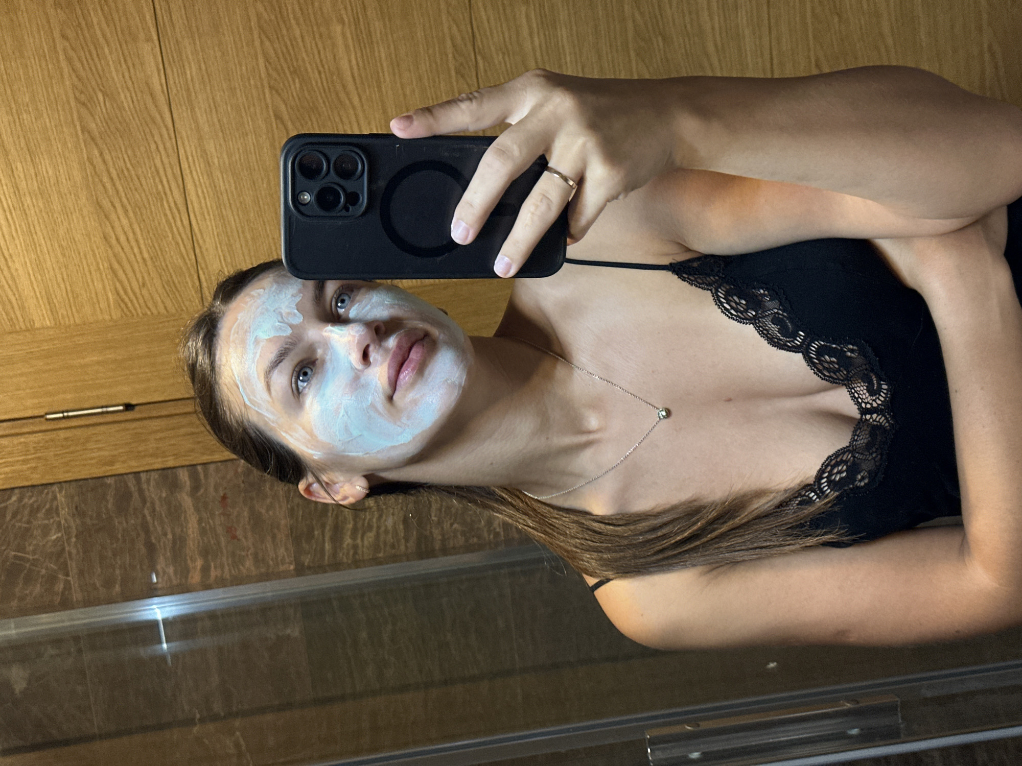 A woman with a light blue face mask applied is taking a selfie in a bathroom. She is wearing a black lace-trimmed camisole. Her hair is pulled back in a ponytail. She is wearing a silver necklace with a small green pendant and a thin silver ring on her right hand. She is holding a black smartphone in her right hand, which is extended to capture the reflection in a mirror. The background shows a shower stall on the left and a wooden paneled wall on the right. The location is identified as Donji Pobori, Montenegro.