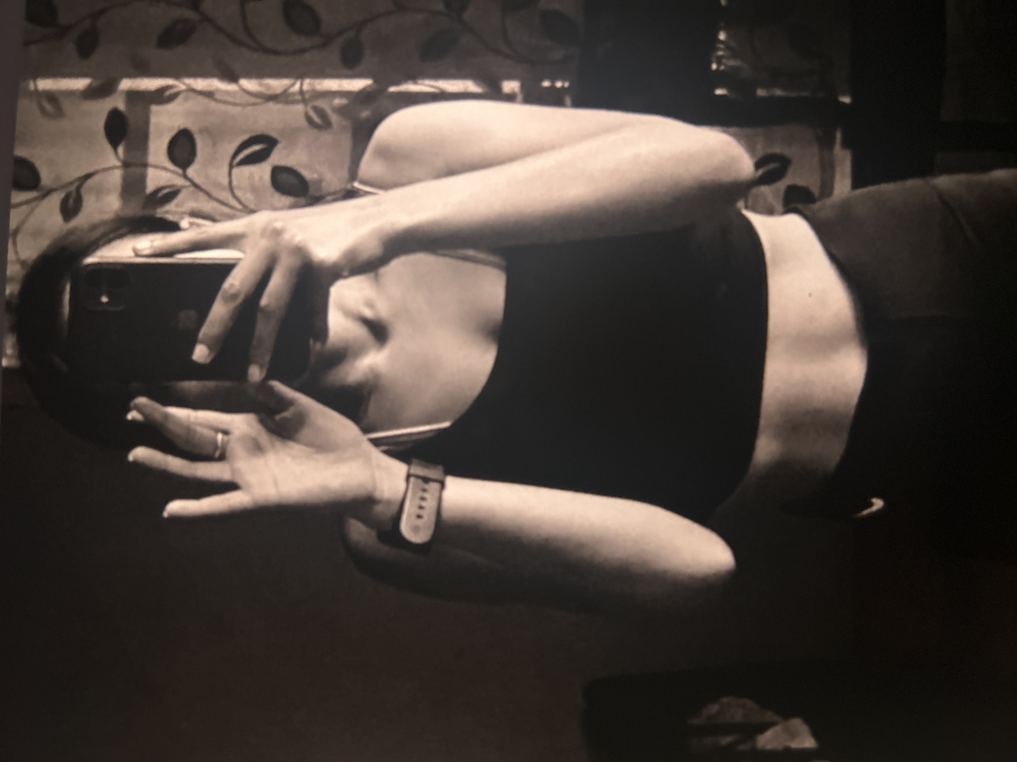 A woman is holding a smartphone and taking a picture. She is wearing a sports bra and leggings, and a watch on her left wrist, and a wedding ring. The location of the image is in a room with floral print curtains. The city and location are unknown.