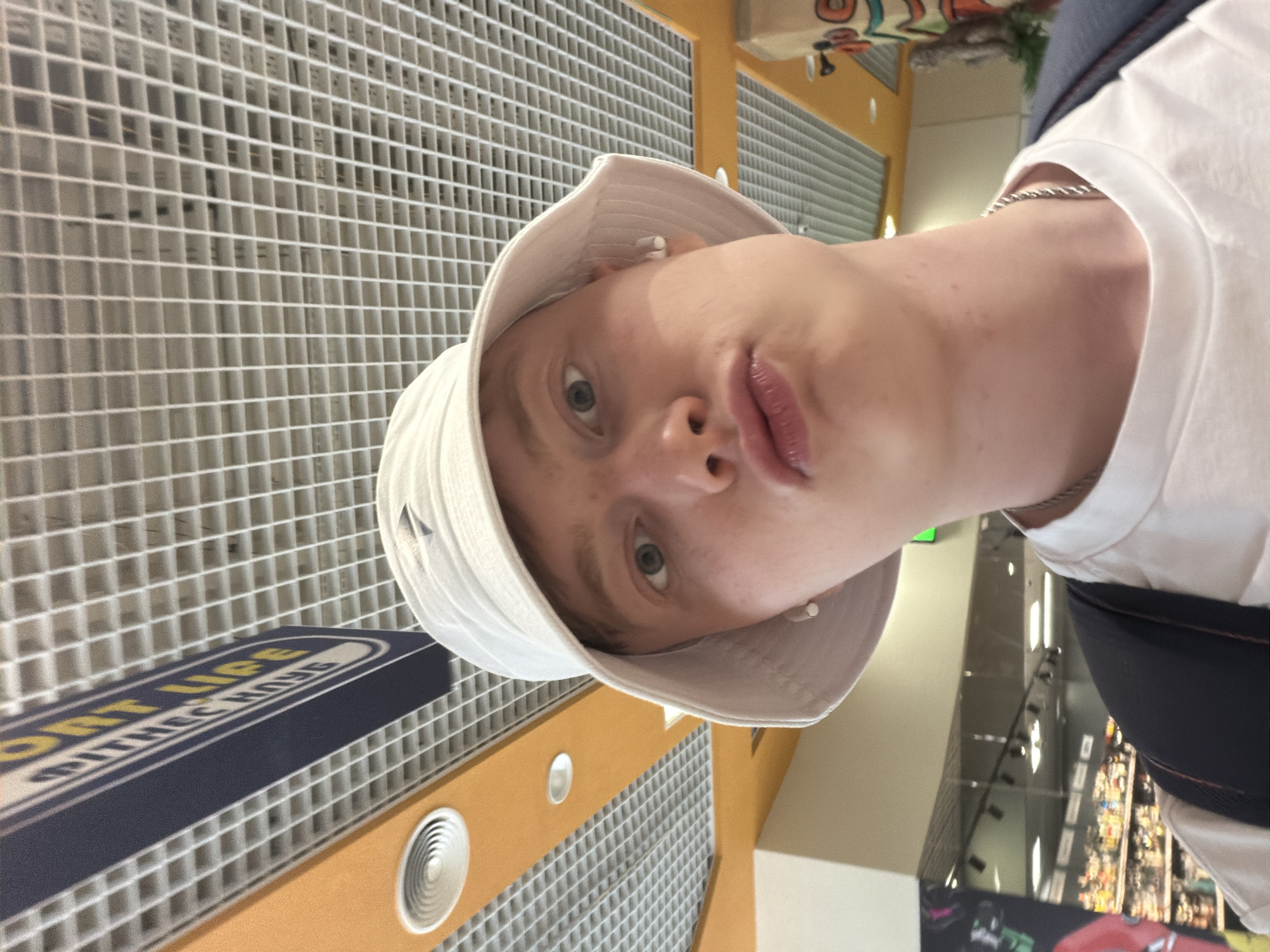 A young man, wearing a white bucket hat and a white t-shirt, is taking a selfie indoors. He has light skin and light-colored hair. He is wearing white earbud. The background shows a grid ceiling, a sign with blue and yellow lettering, and fluorescent lights. In the background, there are shelves stocked with items, suggesting a retail environment like a grocery store. The lighting is bright and even. The photo appears to be taken from a low angle, looking upwards.