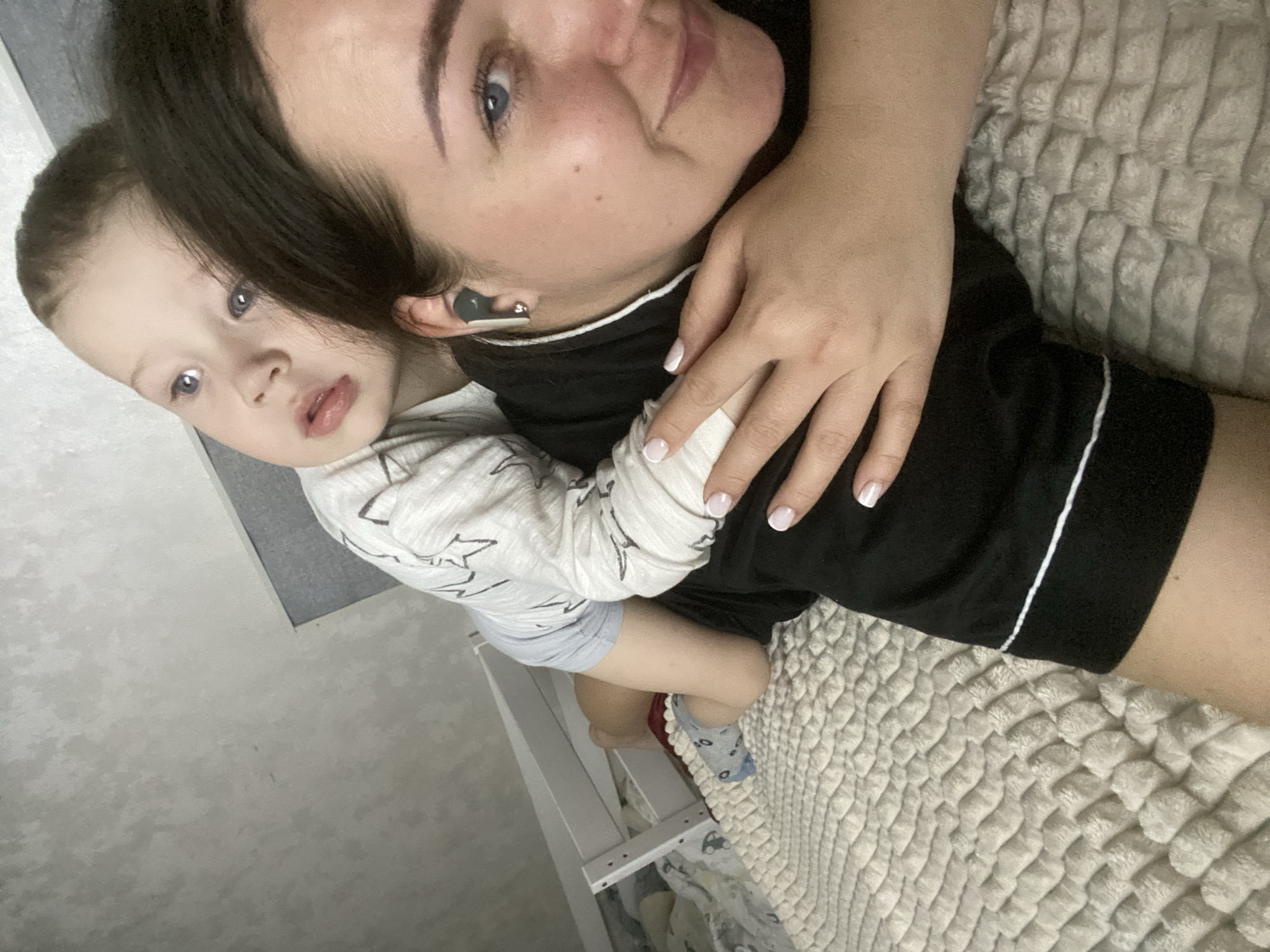 A woman and a young child are lying on a bed covered with a textured, quilted blanket. The woman is positioned at the top of the frame, her head turned slightly to the left. She has dark hair, fair skin, and is wearing a black garment with white trim. An earbud is visible in her left ear. Her hand rests on her upper chest/shoulder area.
The child is situated towards the lower left of the frame, partially obscured by the woman's body. The child has fair skin, light-colored hair, and is wearing a long-sleeved shirt with a print of some kind, light blue shorts, and mismatched socks with a pattern on it.
A bed frame or other structure is visible behind the child. The backdrop includes a light-colored wall.
The location is unknown. The city of Sochi, Russia cannot be determined from this image.