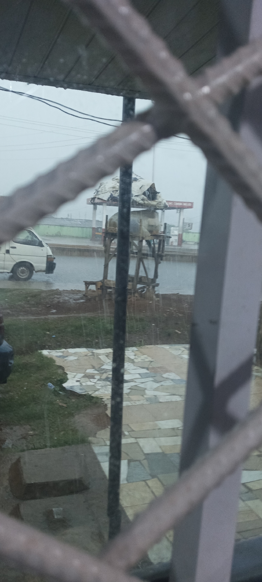 The image, taken from behind a barred window, shows a street scene in the rain. A white van is parked on the left side of the street. A water tower, covered with a white tarp, stands on a wooden platform. Across the street, a building with a red sign is visible, possibly a gas station. In the foreground, a tiled patio or walkway is visible through the bars of the window. The location appears to be Unknown, Unknown.