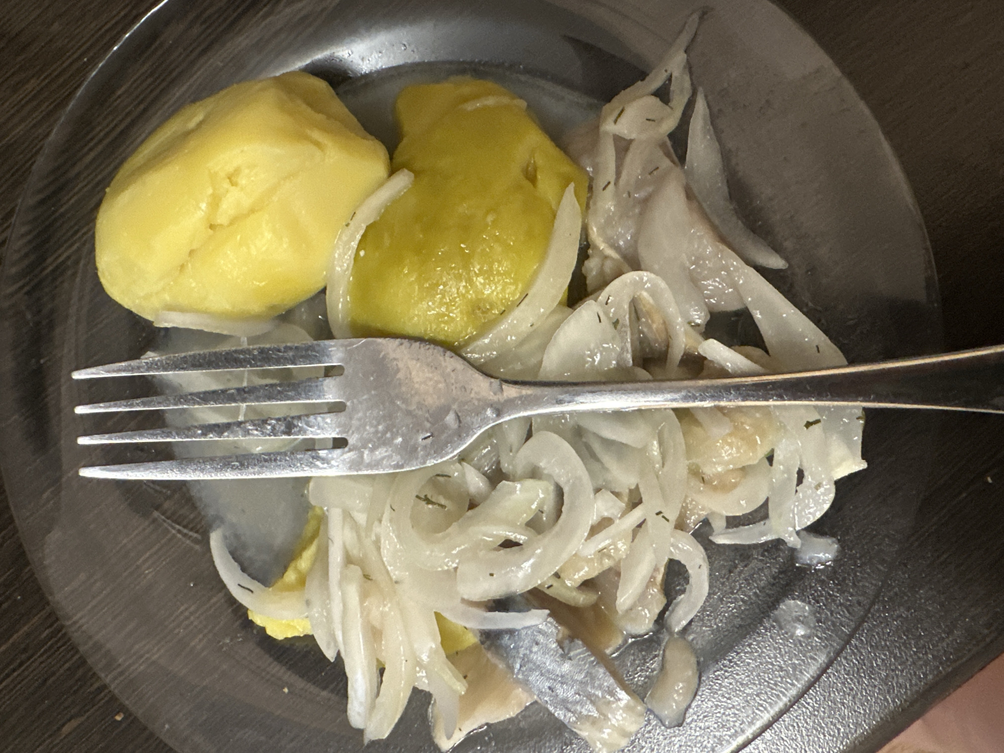 The image shows a plate of food with a fork. The food appears to consist of pickled herring, sliced onions, and what looks like boiled potatoes or dumplings. The setting is indoors, likely a home kitchen or dining area. There is no indication of the location being in Novoaltaysk, Russia. The image is a close-up, top-down view of the plate, with the fork positioned in the center of the food. The lighting is somewhat dim, suggesting it might be evening or taken with artificial light. The plate is made of clear plastic or glass, and it rests on a dark wooden surface. The overall impression is of a simple, home-cooked meal.