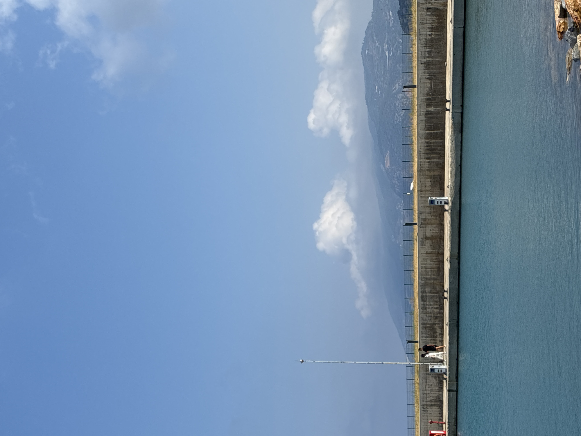 A clear blue sky with scattered white clouds stretches over a harbor in Gazipaşa, Turkey. In the foreground, the tranquil turquoise water of the harbor is visible, with a rocky shore on the right. A concrete pier runs horizontally across the middle ground. On the pier, two figures are walking: a person in a white dress and a person in a black t-shirt and shorts. A tall, thin pole with a light fixture at the top stands near the figures. Beyond the pier, a chain-link fence runs along a grassy area, backed by a concrete wall. In the background, a range of hazy mountains forms the horizon. The scene depicts a peaceful day at the harbor, with the clear sky and calm water suggesting pleasant weather.