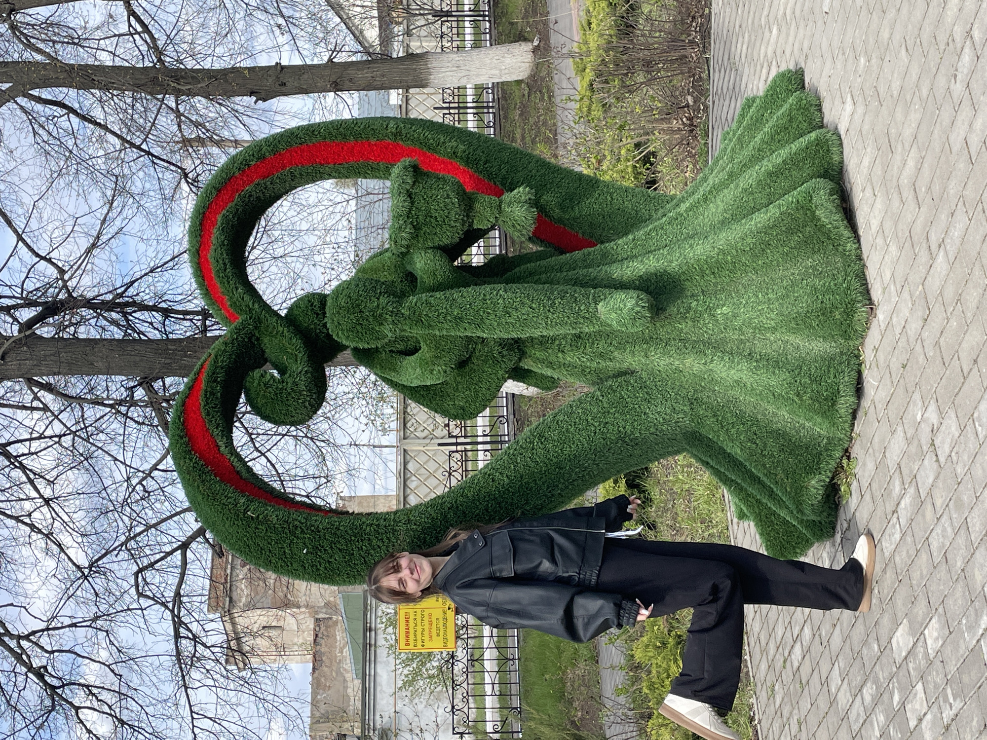 A young woman stands next to a large, green, heart-shaped topiary sculpture with a red stripe on its upper loop. The sculpture depicts a man and a woman in an embrace. The woman wears a black leather jacket, black pants, and white shoes. A yellow warning sign is visible behind the sculpture, attached to a black metal fence. In the background, bare trees reach toward a pale blue sky. The ground is paved with interlocking gray bricks. A building is partially visible in the background, behind the fence. This scene is taking place in the city of Urussu, Russia.