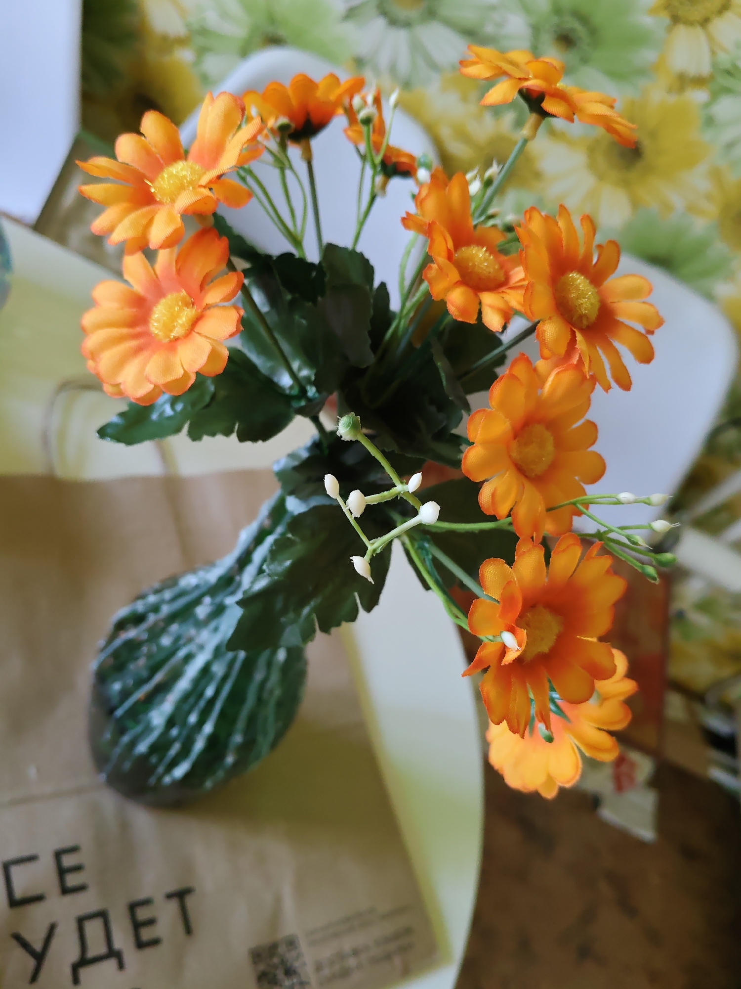 The image displays a close-up, overhead shot of a bouquet of artificial orange daisies with yellow centers, arranged in a dark green, ribbed vase. The vase is placed on a brown paper bag with black text, some of which is legible, reading "СЕ УДЕТ". The background features a blurry floral-patterned wallpaper in shades of green and yellow, and a white surface. The scene appears to be an indoor setting, likely a table or countertop, with soft lighting. There are no people visible in the image. The photo was taken in Rostov-on-Don, Russia.