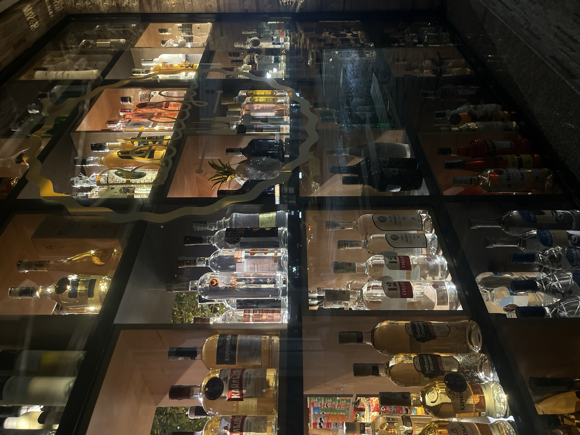 The image presents a view of a lit liquor cabinet, seemingly located in Goce Delcev, Bulgaria, given the Cyrillic labels on some of the bottles. The cabinet is divided into multiple shelves, displaying a variety of liquor bottles arranged horizontally. The composition is shot through a glass panel with decorative etched swirls. The bottles have labels that include "КАРНОБАТСО," "КАЙЛЪШИ," "OUZU," "APER," and "BACA". The light reflects off the glass and bottles, creating a distorted view.