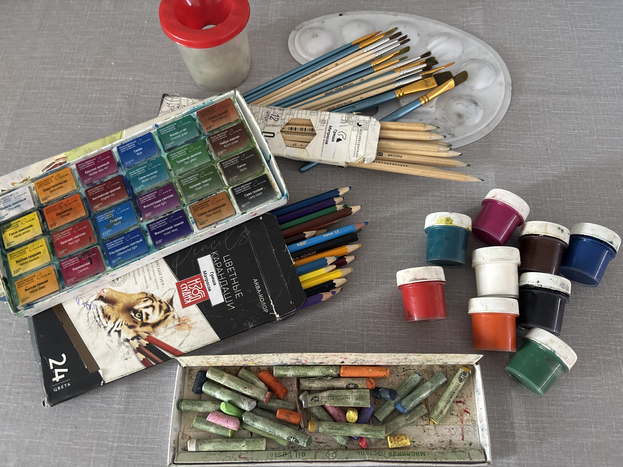 The image depicts a collection of art supplies laid out on a gray surface.
**Objects:**
* An open watercolor paint set labeled "Sonet" with a tiger illustration on the box.
* A partially open box of colored pencils labeled "Grisha Matrosov".
* A container of oil pastels.
* Several small jars of paint with white lids.
* A collection of paintbrushes in a cardboard box.
* A plastic paint palette.
* A red-rimmed water container.
**Text on objects:** The text is mainly in Russian. The watercolor set has individual color labels in Russian and English (e.g., "Zinc white", "Gold ochre"). The colored pencil box is labeled with "цветные карандаши", which translates to "colored pencils". The watercolor box says "Невская палитра" "Nevskaya Palitra" - “The Nevskaya Palitra” Art Materials Factory is a well-known art supplies manufacturer, located in Saint Petersburg, Russia.
The colored pencil box says "Турский Тигр".
The pastels are also labeled with "Невская палитра".
**Overall Impression:** The image seems to be a collection of a person's art supplies used for painting and drawing.