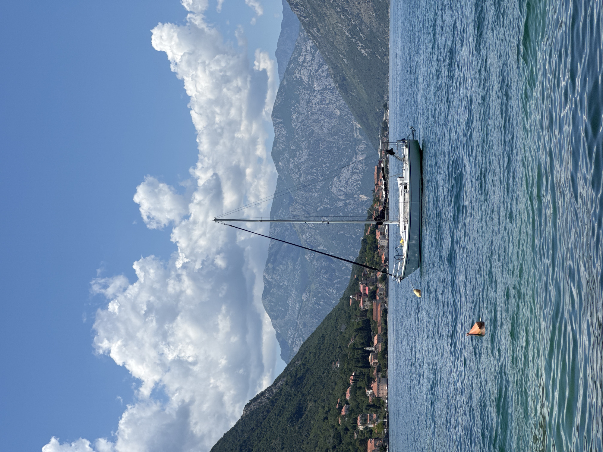 The image depicts a serene coastal landscape in Dobrota, Montenegro, under a partly cloudy blue sky. A white sailboat, its mast upright and sails furled, is anchored in the mid-ground of a large body of blue-green water. The boat's bow faces slightly towards the left. A small yellow buoy is tethered to its front. In the foreground, an orange buoy floats on the rippled water. To the left, a steep, densely vegetated mountain slope descends towards the water, where a cluster of buildings with terracotta roofs forms a village. A prominent white church tower or spire rises among the structures. In the background, across the expanse of water, additional rugged, grey-blue mountains frame the scene. No individuals or active movements are visible.