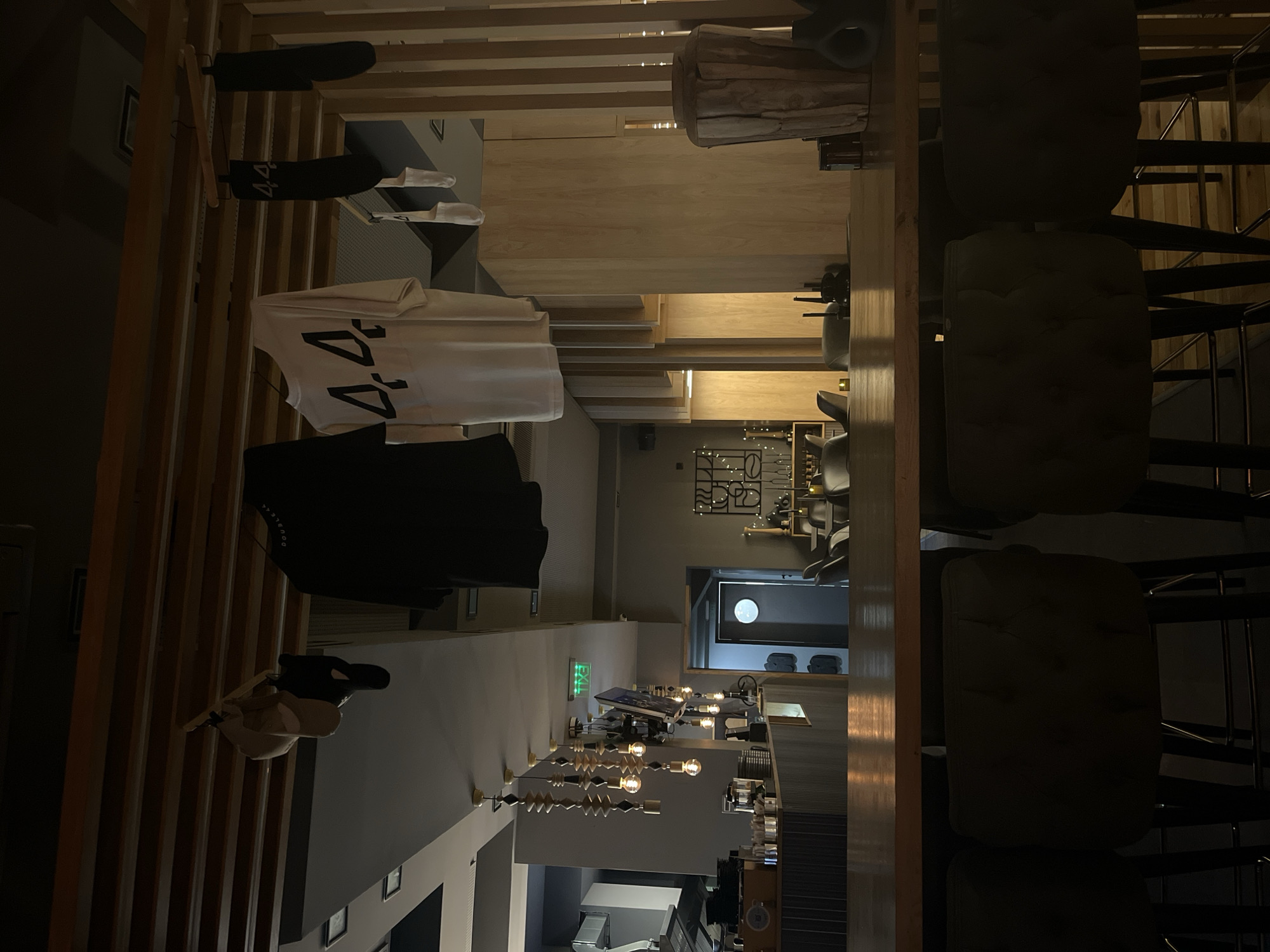 The image depicts the interior of a commercial space, likely a retail store or restaurant, in Varna, Bulgaria. The foreground features vertical wooden slats. On the left, clothing items, including a white shirt with the number "44" on it and a black jacket, are hung. The space continues into the background, showcasing a seating area with chairs, overhead lighting fixtures, and a back room with a circular window. The environment is modern, with gray tones in the walls and floors. There are no people visible in the main area.