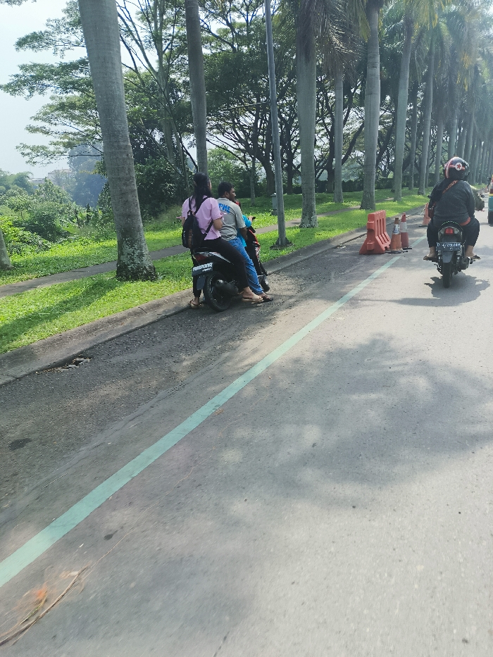 A paved road with a green line down the middle. Two people are on a motorcycle, traveling left to right. The motorcycle is black and has a white license plate. There is a second motorcycle in the distance, traveling in the same direction, also on the right side of the road. The road is lined with palm trees on the right side. There is a grassy area on the left side of the road, and in the background there are trees and buildings. The location is Padalarang, Indonesia (latitude: -6.853, longitude: 107.492).