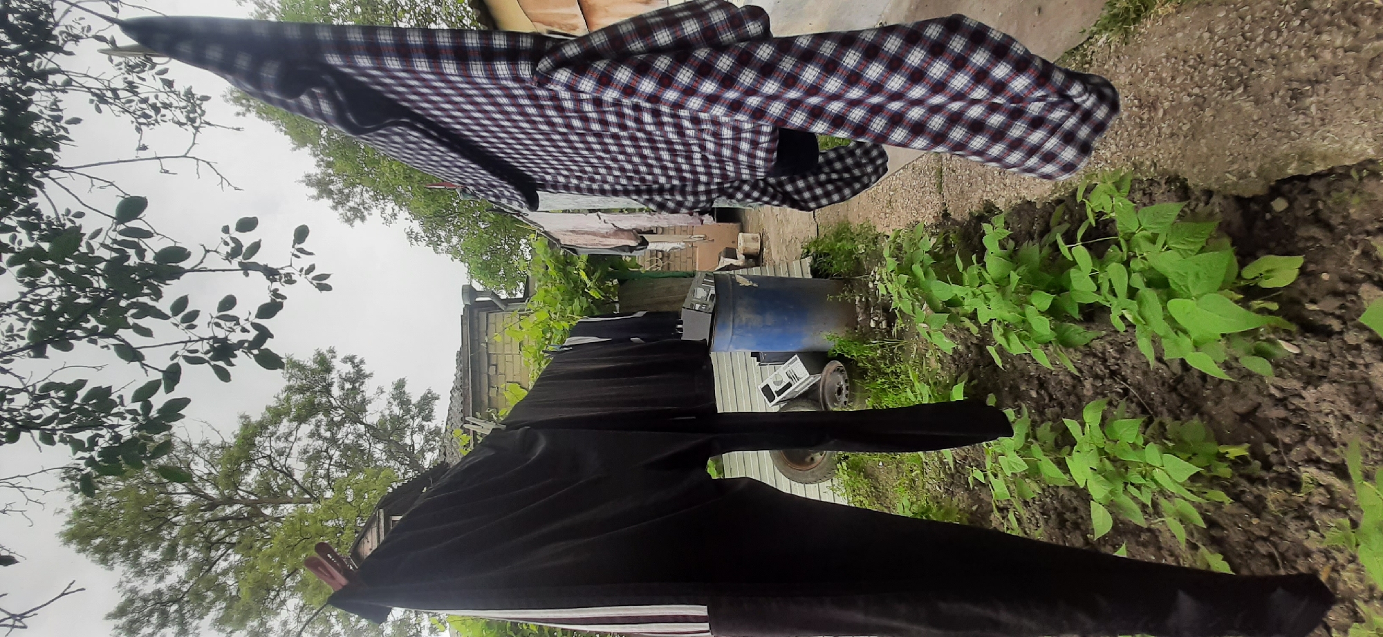 The image depicts an outdoor scene in Yessentukskaya, Russia, likely a backyard or a communal area. Laundry is hanging on a clothesline, including a pair of black pants with white stripes and a red and white checkered shirt. Various objects are scattered in the background, such as a blue barrel, car tires, and a small building with a pitched roof. The area is overgrown with green plants, suggesting a rural or semi-rural setting. The sky is overcast, indicating potentially cloudy weather. No people are visible in the image.