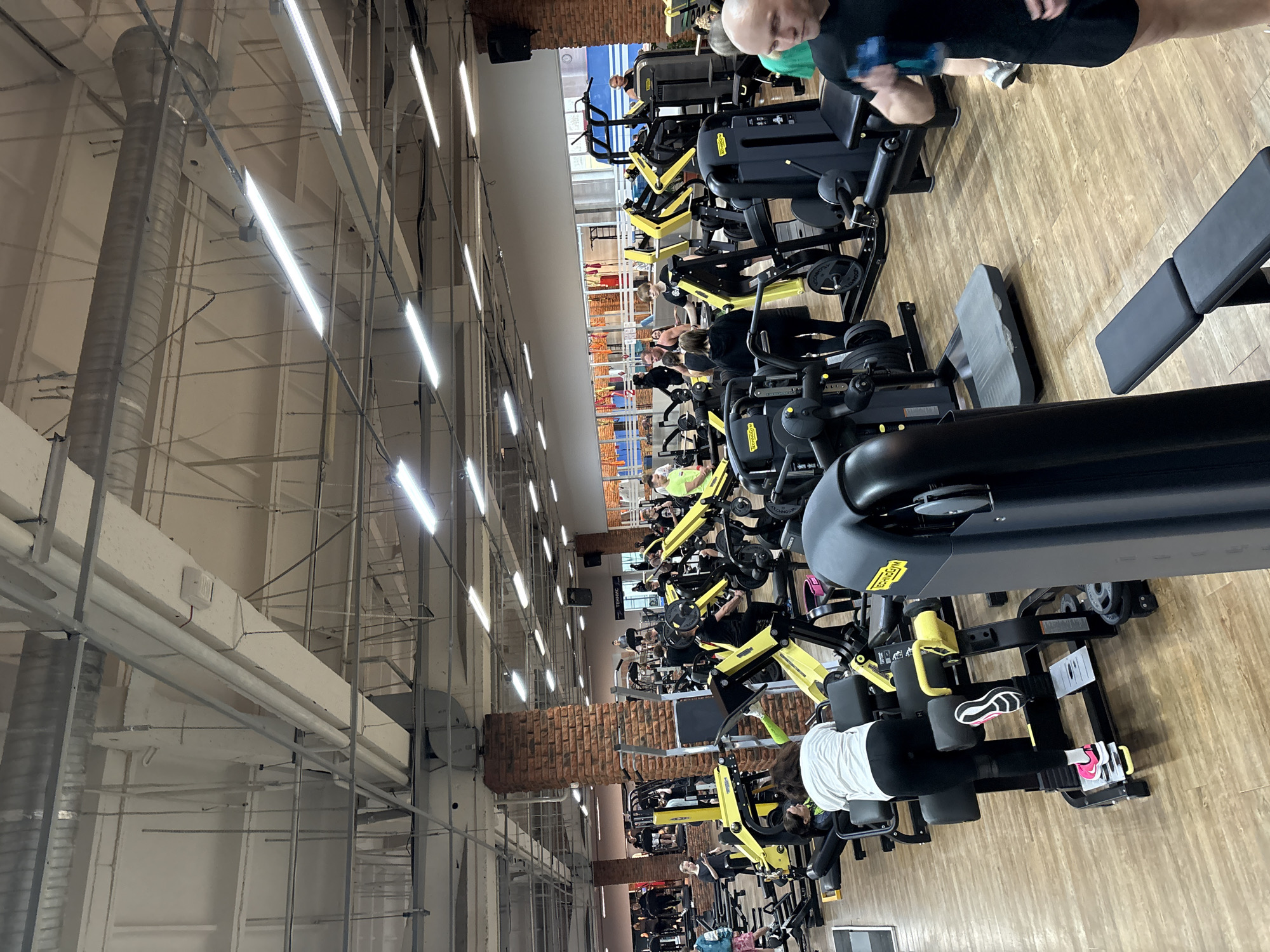 The image shows the interior of a gym in Chaikovsky, Russia. A row of weight machines, mostly black and yellow in color, lines the right side of the frame, with people using them. The gym has a high ceiling with exposed ductwork and fluorescent lighting. A brick wall section is visible further back. The floor appears to be wood or laminate. Several individuals are actively using the equipment. A man with a shaved head is visible at the upper right of the frame.