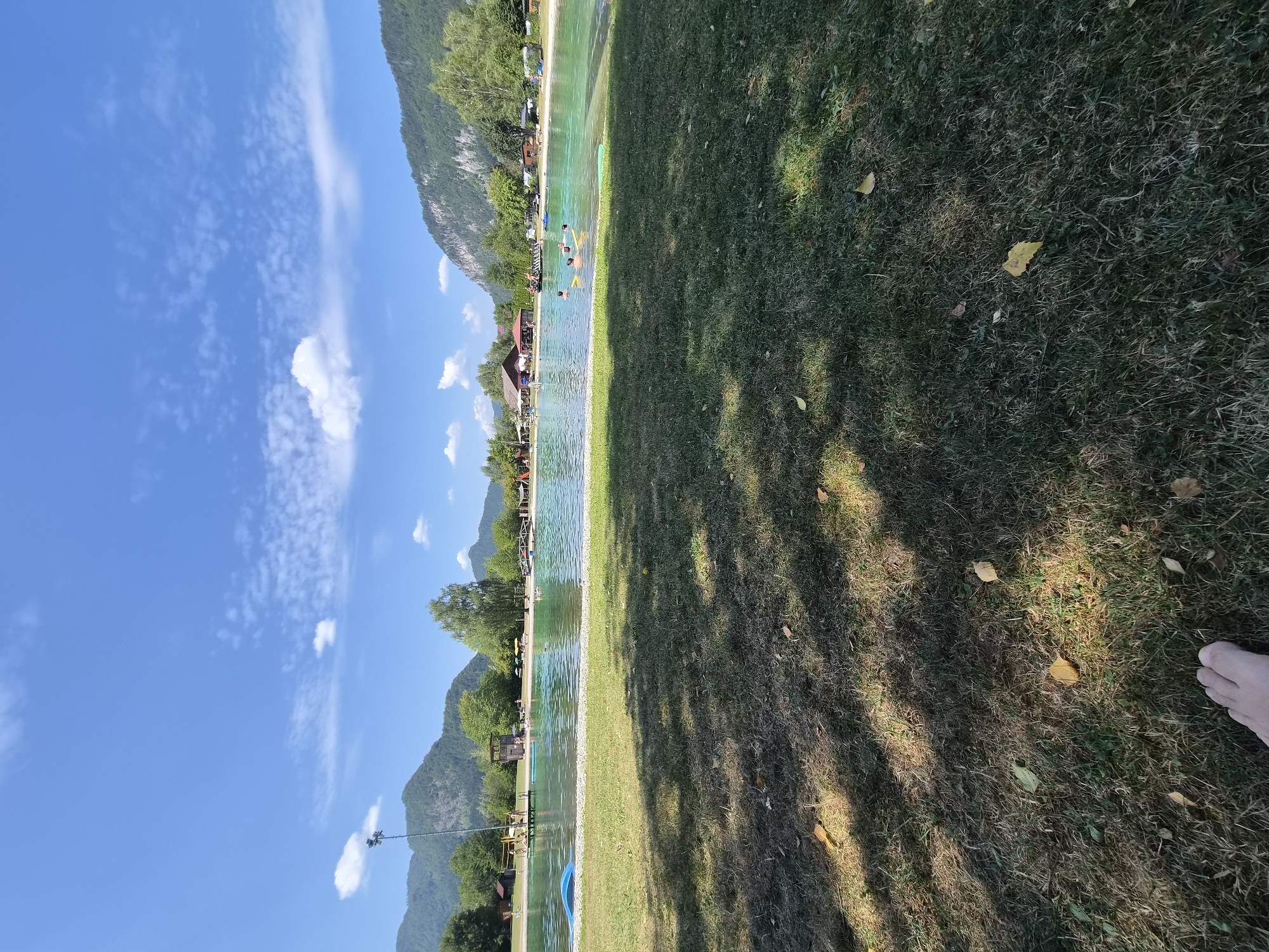The image depicts an outdoor swimming area on a sunny day, likely in Landl, Austria, given the mountainous backdrop and lush green surroundings. The foreground is dominated by a grassy area with scattered fallen leaves and shadows cast by trees, with a bare foot visible in the lower left corner. Beyond the grass, a large, clear blue swimming lake or pool is the central focus. Several people are visible in the water, some swimming and others relaxing. The lake is bordered by a light-colored shore and then more greenery. In the background, various structures can be seen, including buildings that appear to be part of a recreational facility, a lifeguard tower, and what might be changing rooms or pavilions. The scene is framed by green trees and imposing mountains under a bright blue sky dotted with white, wispy clouds.