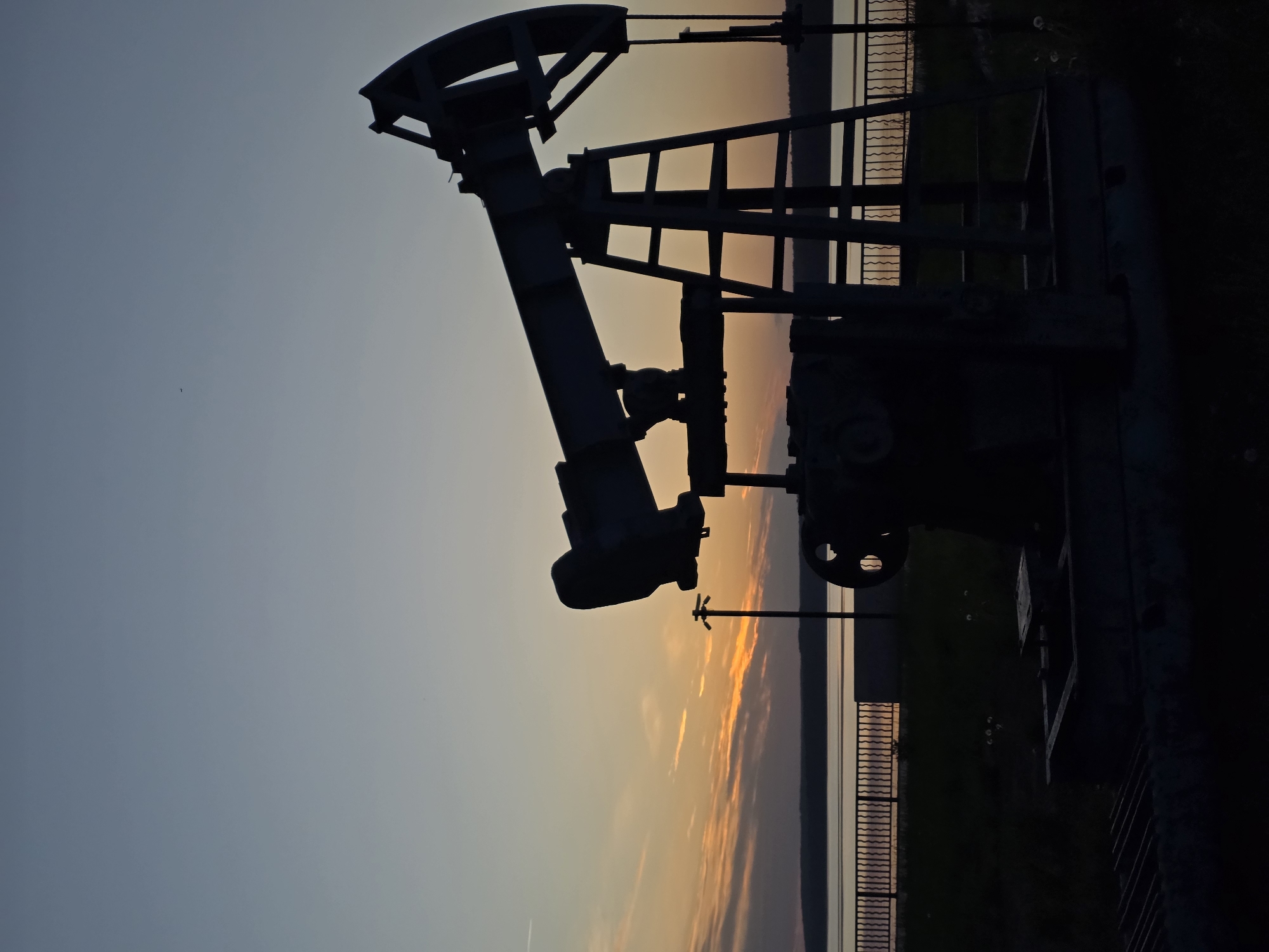 The image depicts an oil pump jack silhouette against a sunset sky in Chaikovsky, Russia. The pump jack is positioned to the left of the frame, obscuring a view of a body of water and adjacent land behind a fence. The foreground shows an embankment wall.