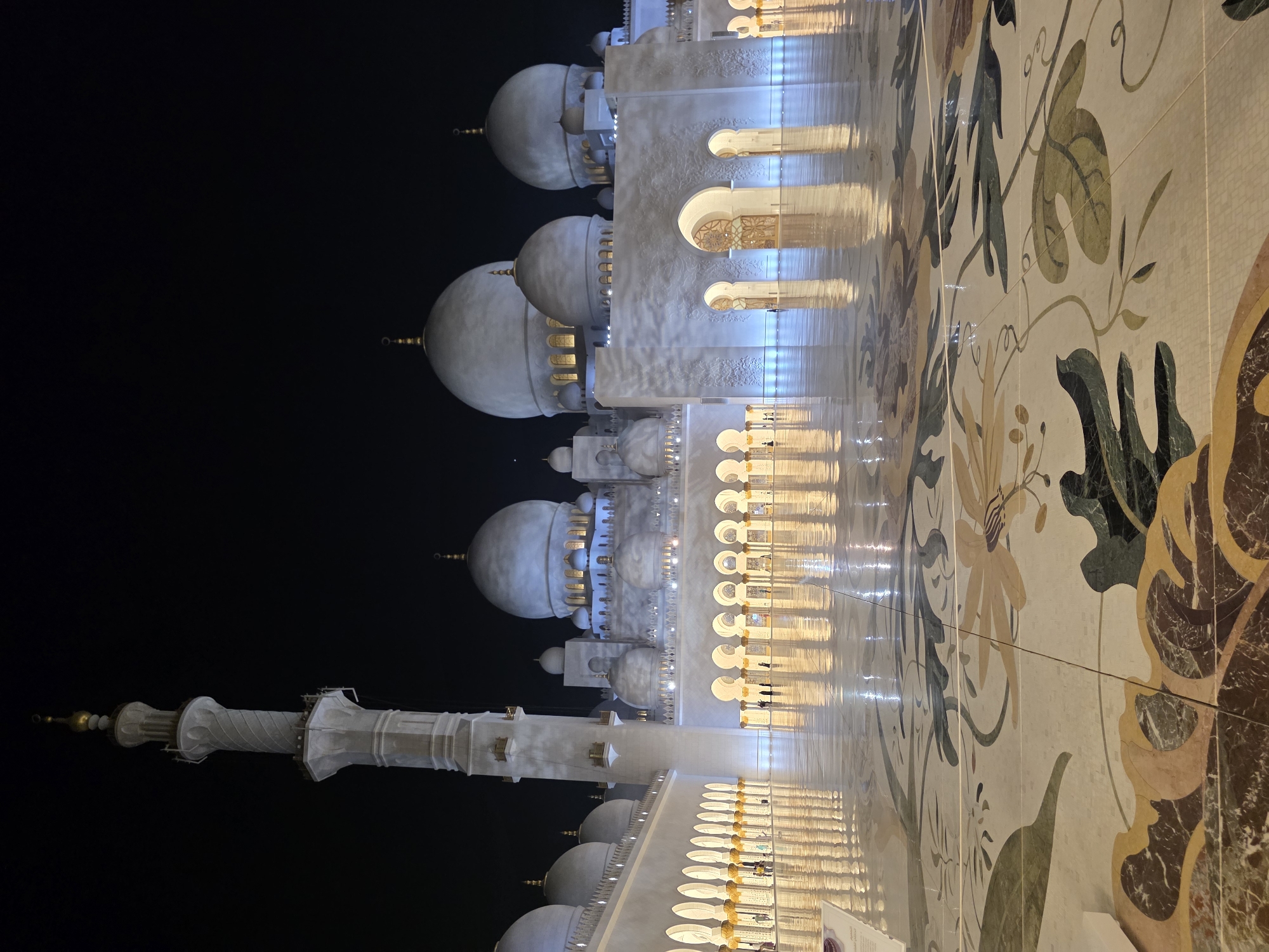 The image captures the Sheikh Zayed Grand Mosque in Abu Dhabi, United Arab Emirates, at night. The grand courtyard is illuminated, with its polished marble floor reflecting the surrounding architecture. The floor is decorated with intricate floral mosaics in various earthy and green tones, bordered by darker marble.
The mosque's exterior is a prominent feature, with its white marble walls and numerous domes, domes, and minarets. The central courtyard is lined with arcades, each supported by columns adorned with elaborate details. The lighting casts a warm glow on the intricate carvings and decorative elements of the building. The sky above is dark, emphasizing the illuminated structure.
A few individuals are visible in the distance, walking along the arcades, adding a sense of scale to the vastness of the mosque. Their presence is minimal, highlighting the grandeur and serene atmosphere of the location. The overall scene depicts a magnificent architectural masterpiece bathed in the tranquil light of the night.