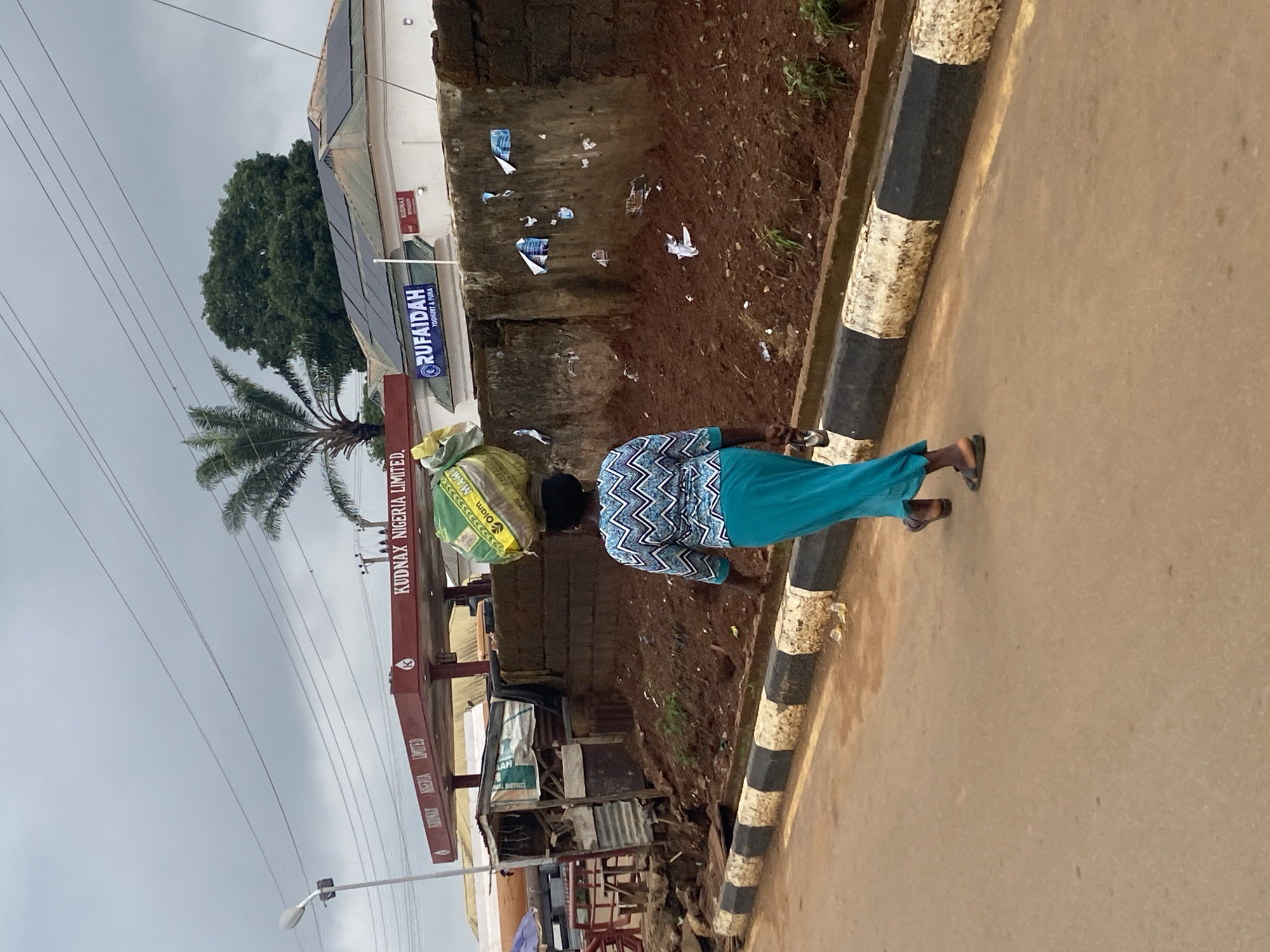 A person is walking along a street next to a curb with alternating black and white markings. They are carrying a green and yellow bag on their head. A building with the sign "RUFAIDAH" can be seen in the background, along with the sign "KUDNAX NIGERIA LIMITED". The surrounding environment includes a palm tree and other structures. The sky is overcast. Based on the signs and other contextual clues in the image, the location is likely Kafanchan B, Nigeria.