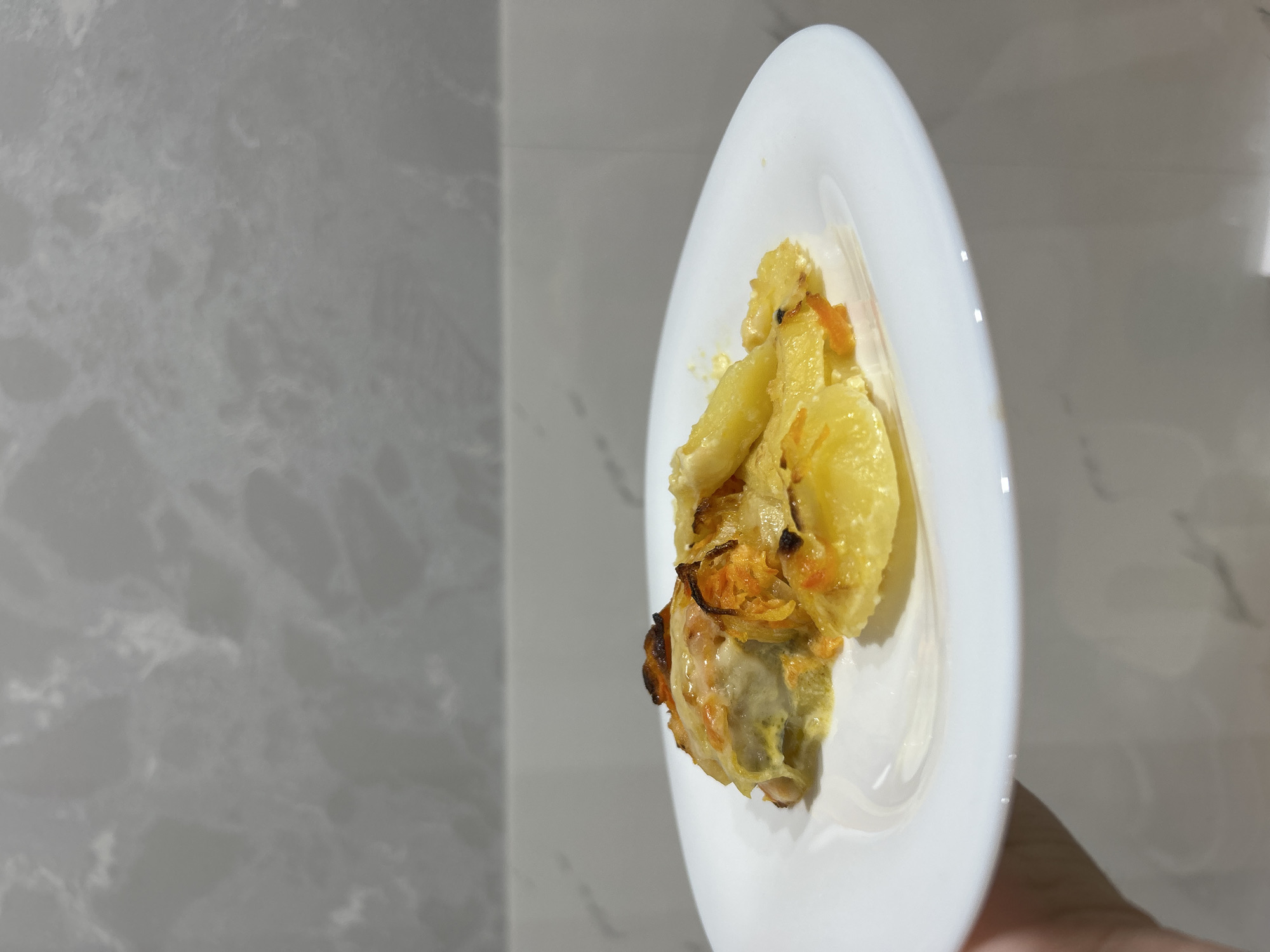 The image shows a plate of food, possibly in Chaikovsky, Russia, though this cannot be confirmed from the image alone. The food appears to be cooked and includes items that appear to be potatoes and other vegetables. The plate is white and has a simple design. The background consists of what appears to be a wall, possibly made of marble or a similar material. A small portion of a hand is visible at the bottom of the frame.