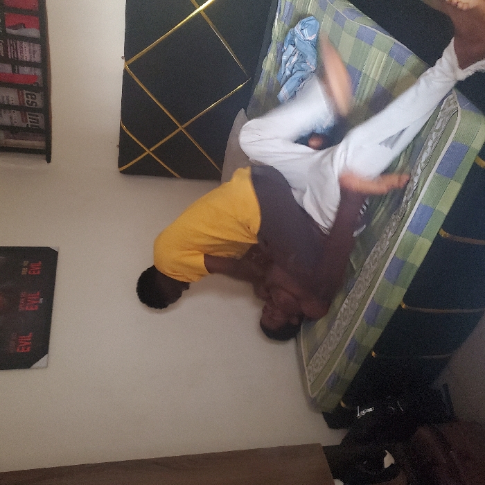 This image captures an interior scene in Eziobodo, Nigeria. Two individuals are on a bed with a black headboard featuring a gold geometric line pattern and a mattress covered by a light green and blue plaid sheet.
One individual, wearing a yellow t-shirt and dark trousers, is positioned kneeling or crouching over another individual. The second individual is shirtless, wearing white trousers, and is lying supine on the mattress. The person in the yellow t-shirt is leaning forward, with hands placed on or near the chest and upper abdomen area of the individual beneath. The lower individual's legs are elevated and display motion blur, indicating recent or ongoing movement. A crumpled light blue garment rests on the mattress near the lower individual's legs.
The background consists of a white wall. A black poster with red text, including phrases like "SEE NO EVIL," is affixed to the wall. Above the headboard, a shelf holds several books or magazines, one clearly displaying "BURSA" on its spine. To the left of the bed, a dark, indistinct object, possibly furniture, is visible.