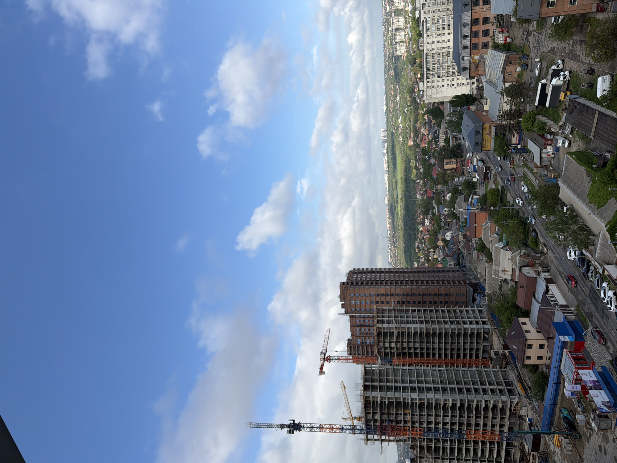 The image shows a cityscape, dominated by ongoing construction of multiple high-rise buildings. Numerous cranes are visible amidst the construction sites. In the background, a mix of residential areas and buildings stretches into the distance. A road runs through the urban landscape. The sky is partly cloudy. While the image likely depicts a construction site in an urban area, there is no definitive evidence provided in this image to confirm its location in Ovoshchnoy, Russia.