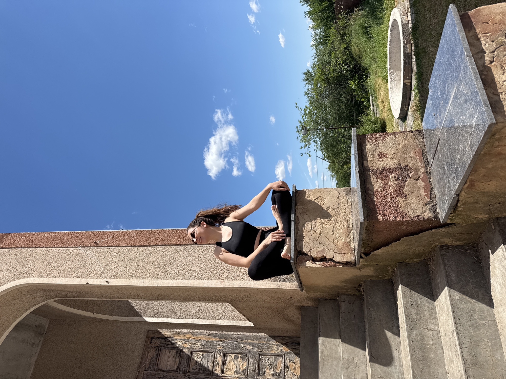A woman in black yoga attire sits cross-legged on a stone ledge outside an arched building with a wooden door. She is looking up and to her right, wearing sunglasses. The building has rough, textured walls and is set against a bright blue sky with scattered white clouds. To the right of the building, there is green foliage and a circular concrete structure in the grass. Stone steps lead up to the ledge on the left side of the frame. The scene appears to be outdoors on a sunny day, possibly in a rural or historical setting, and is situated in the city of Lozuvatka, Ukraine.
